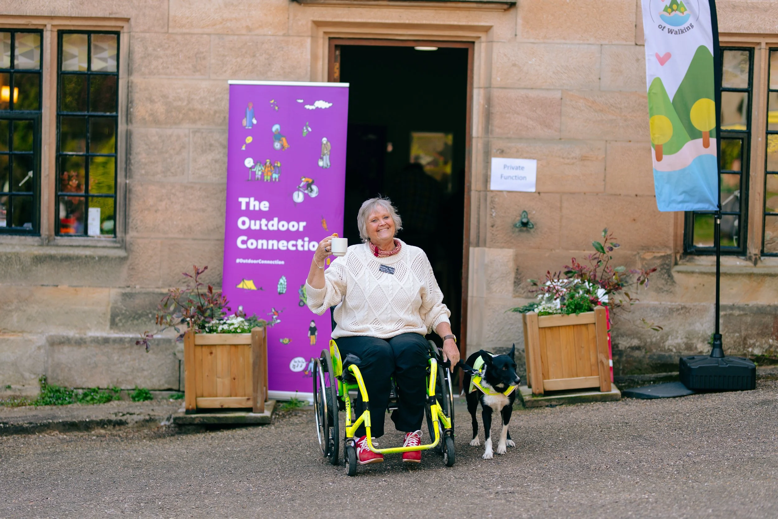 A woman in a wheel chair with a dog smiles in front of a banner that reads The Outdoor Connection