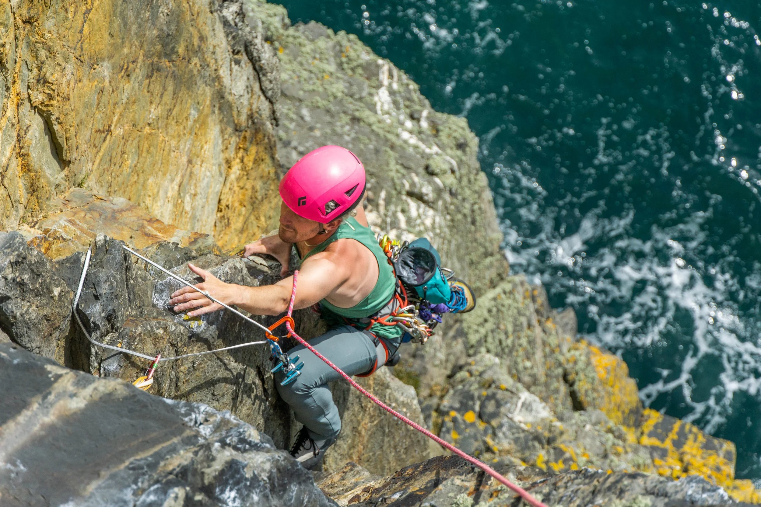 AJ climbs a trad route on a sea cliff. The photo is taken from above and you can see them climbing towards a sling with the sea far below.