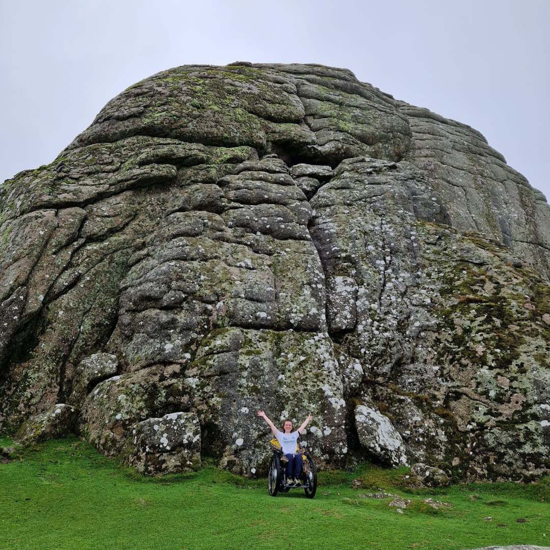  Bailey, who is white and femme presenting, in their all terrain wheelchair posing with hands in the air in front of Haytor Rocks in Dartmoor National  Park 
