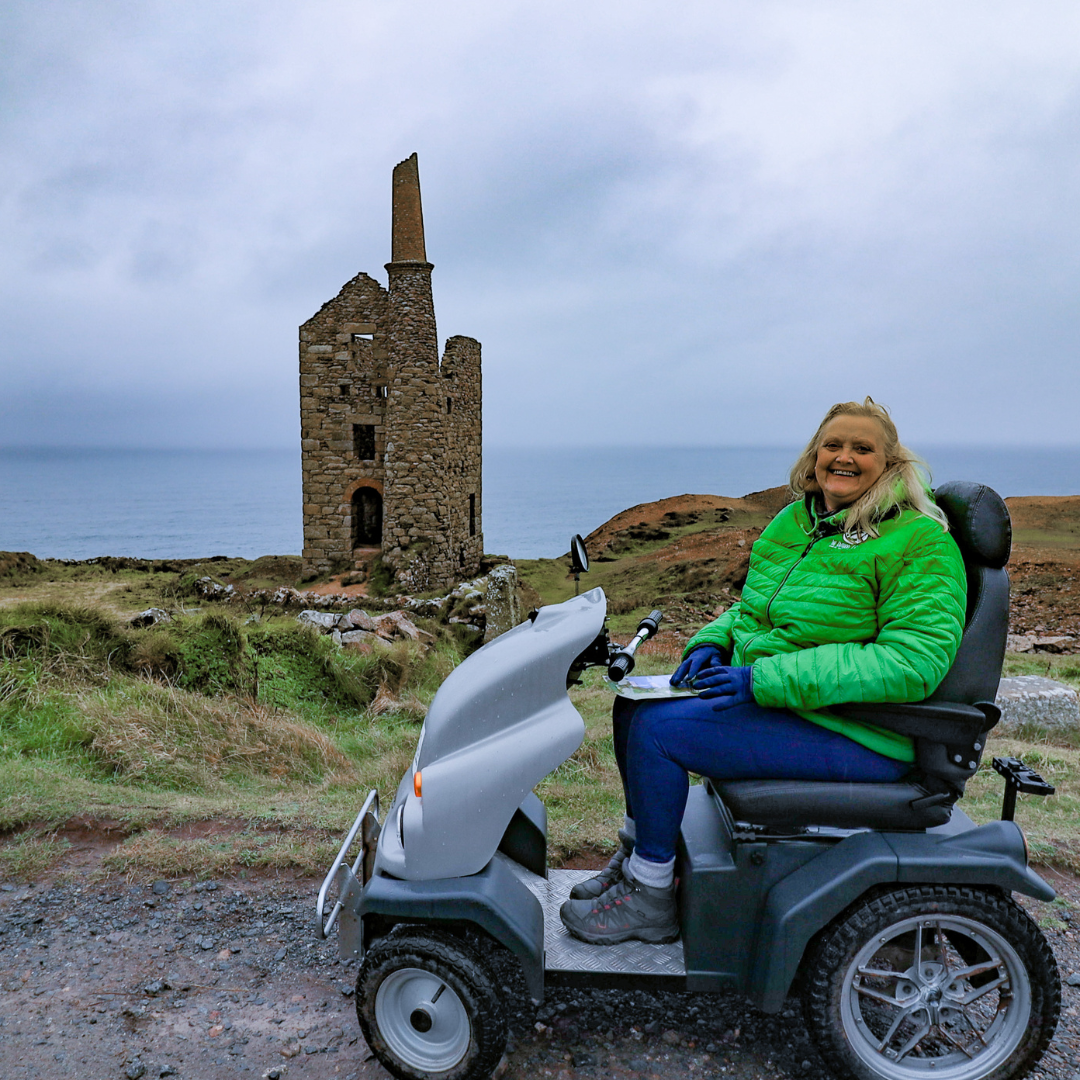  Debbie on their all-terrain mobility scooter outside on a track in front of a large ruin. 