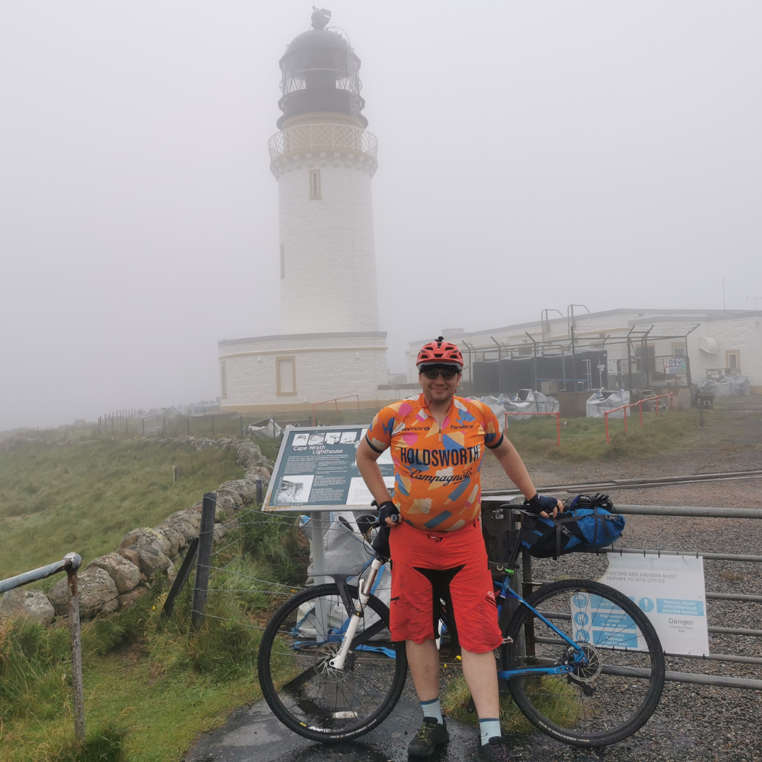  Spencer stood in front of his bike in a cycling gear. It’s foggy and there is a light house behind him. 