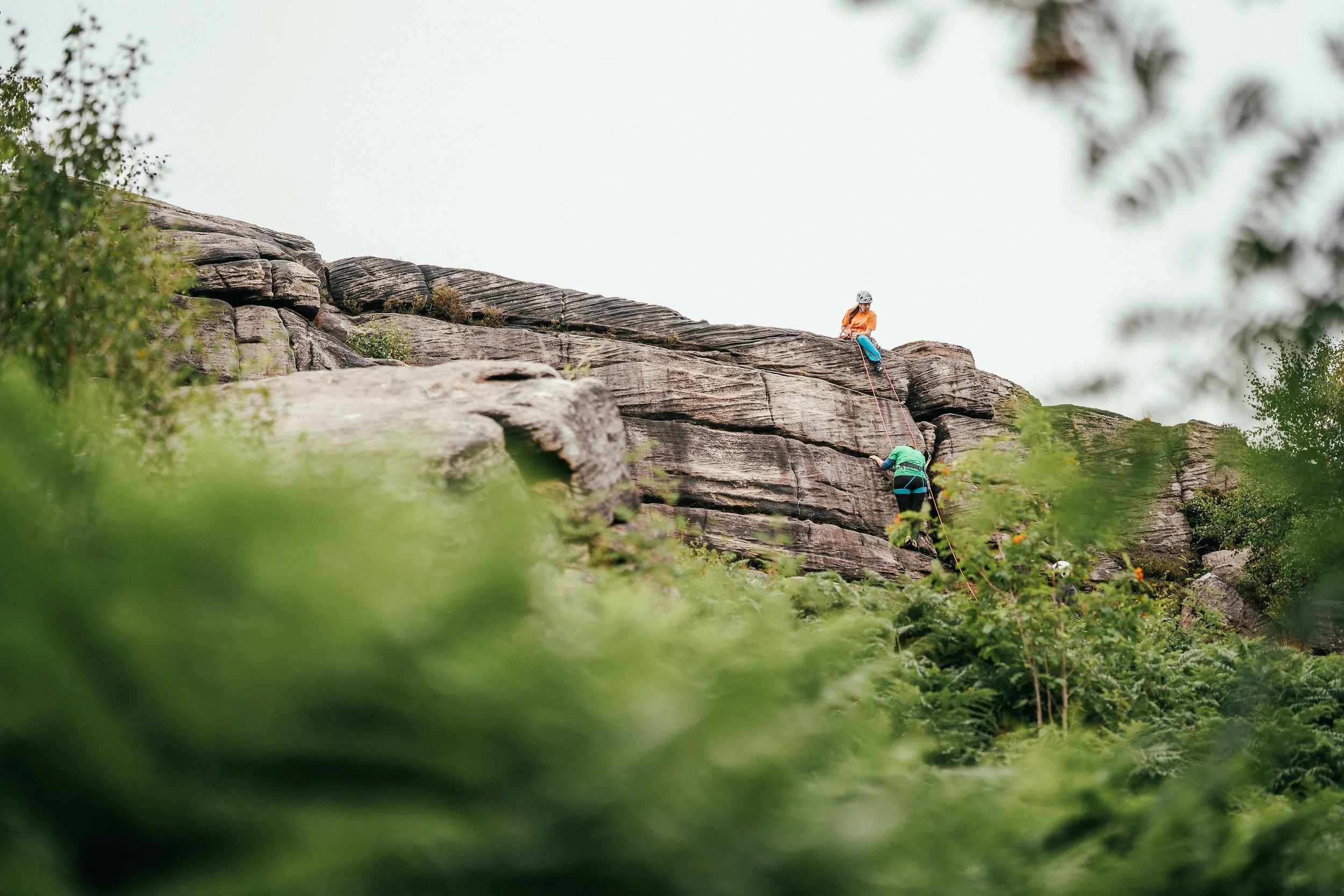 Two women on a trad climb, one sits at the top belaying, the other climbs up. there is blurry green trees in the foreground