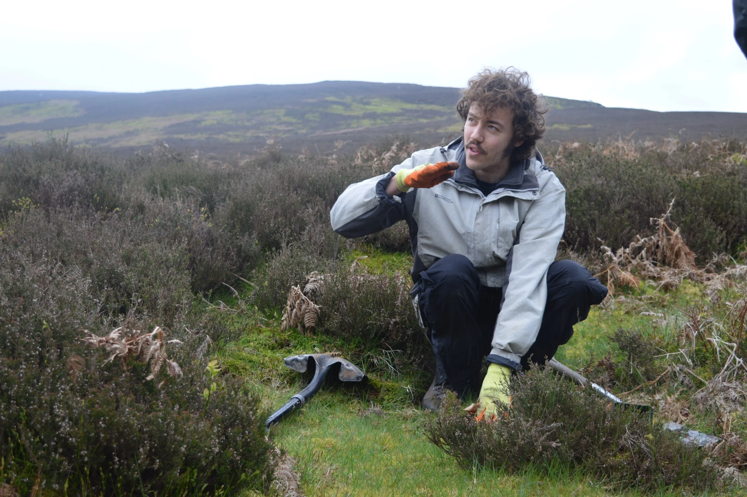 Jack, bends down in the heather, he wears a grey jacket and orange gloves and has a spade next to him. He is white with brown hair and a moustache