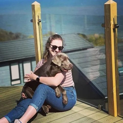 Sam sits holding a chocolate lab with the sea in the background
