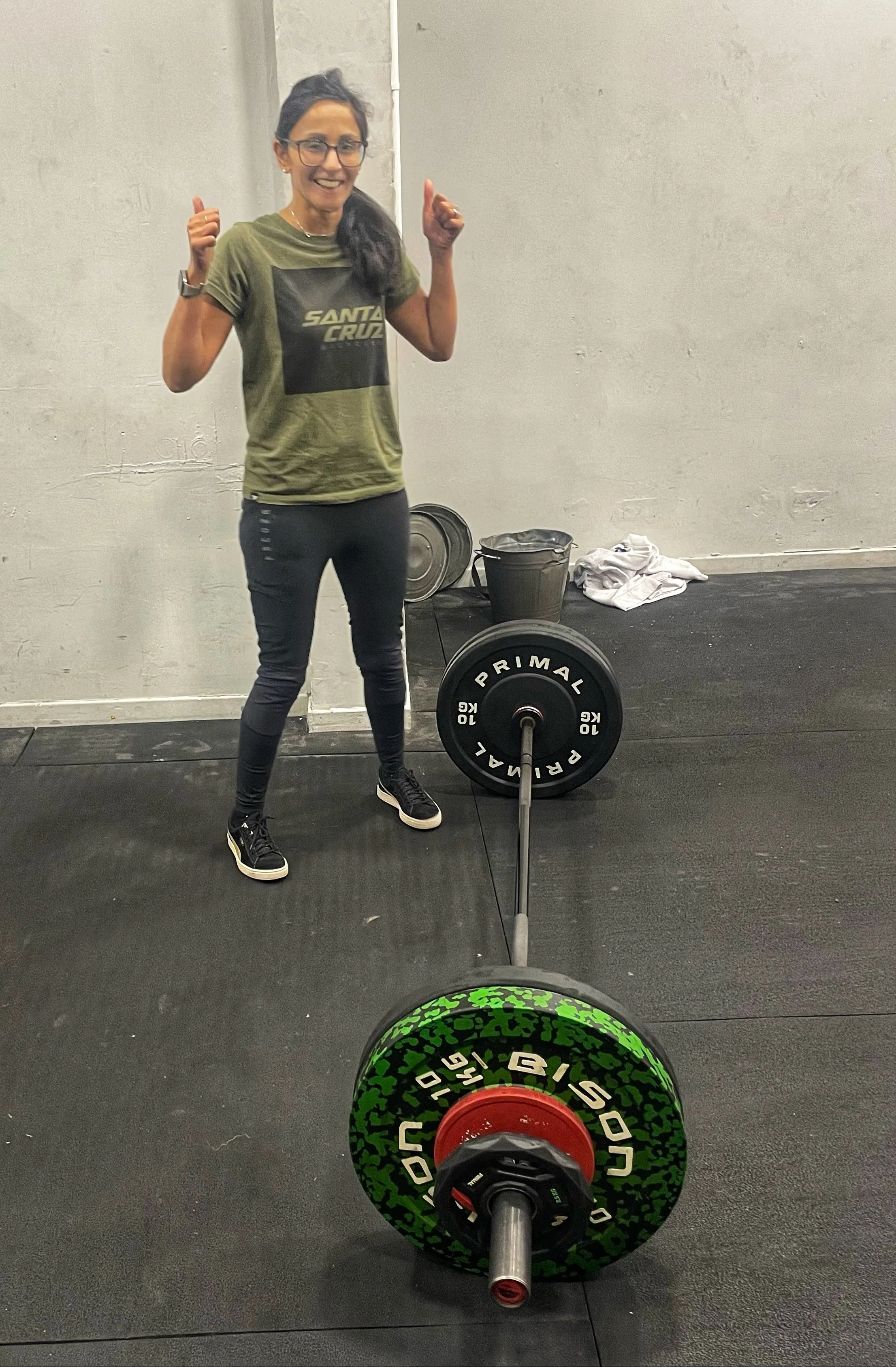 Aneela stands next to a bar with huge weights on it and gives a thumbs up