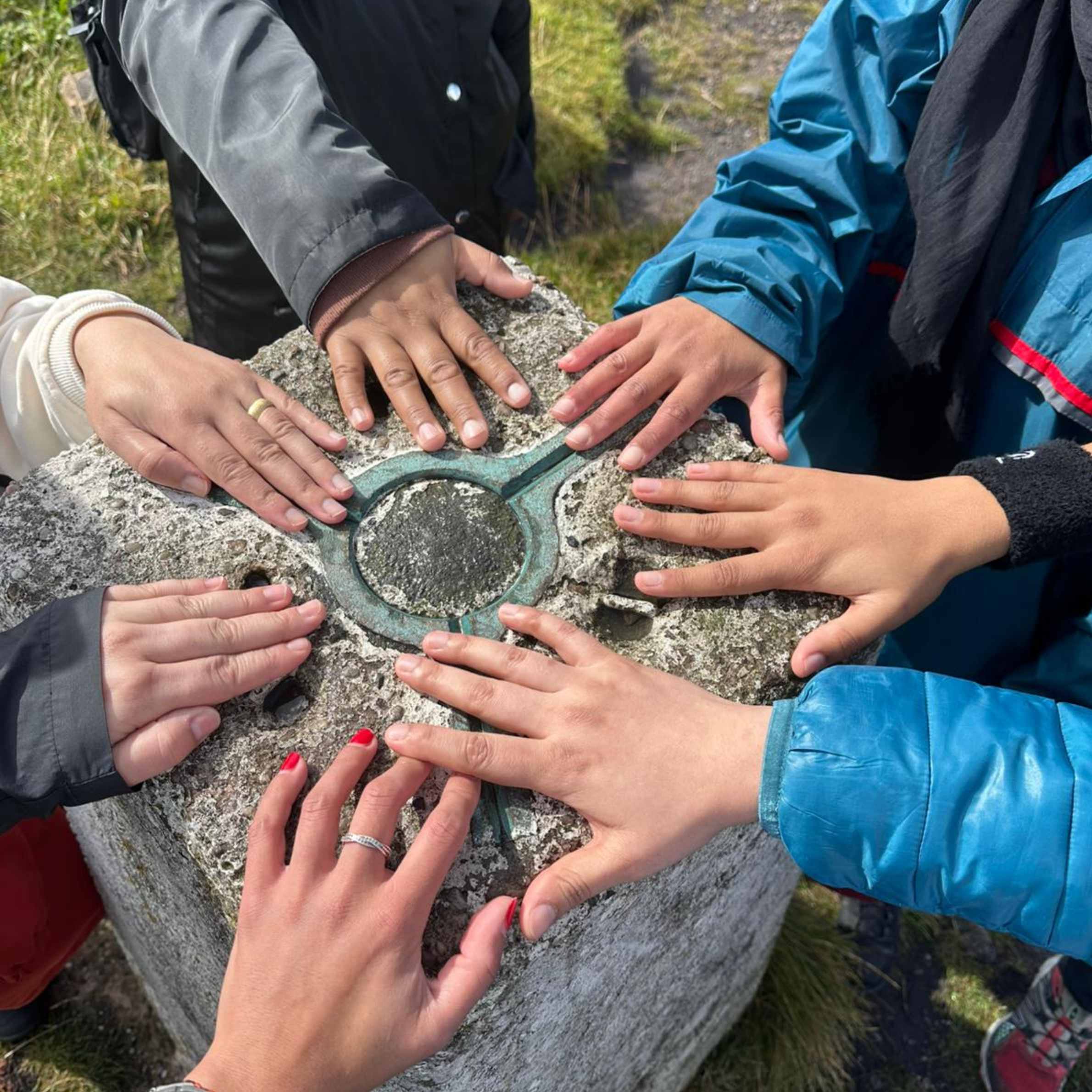 Lots of people's hands touching the top of a trig point, all in a circle