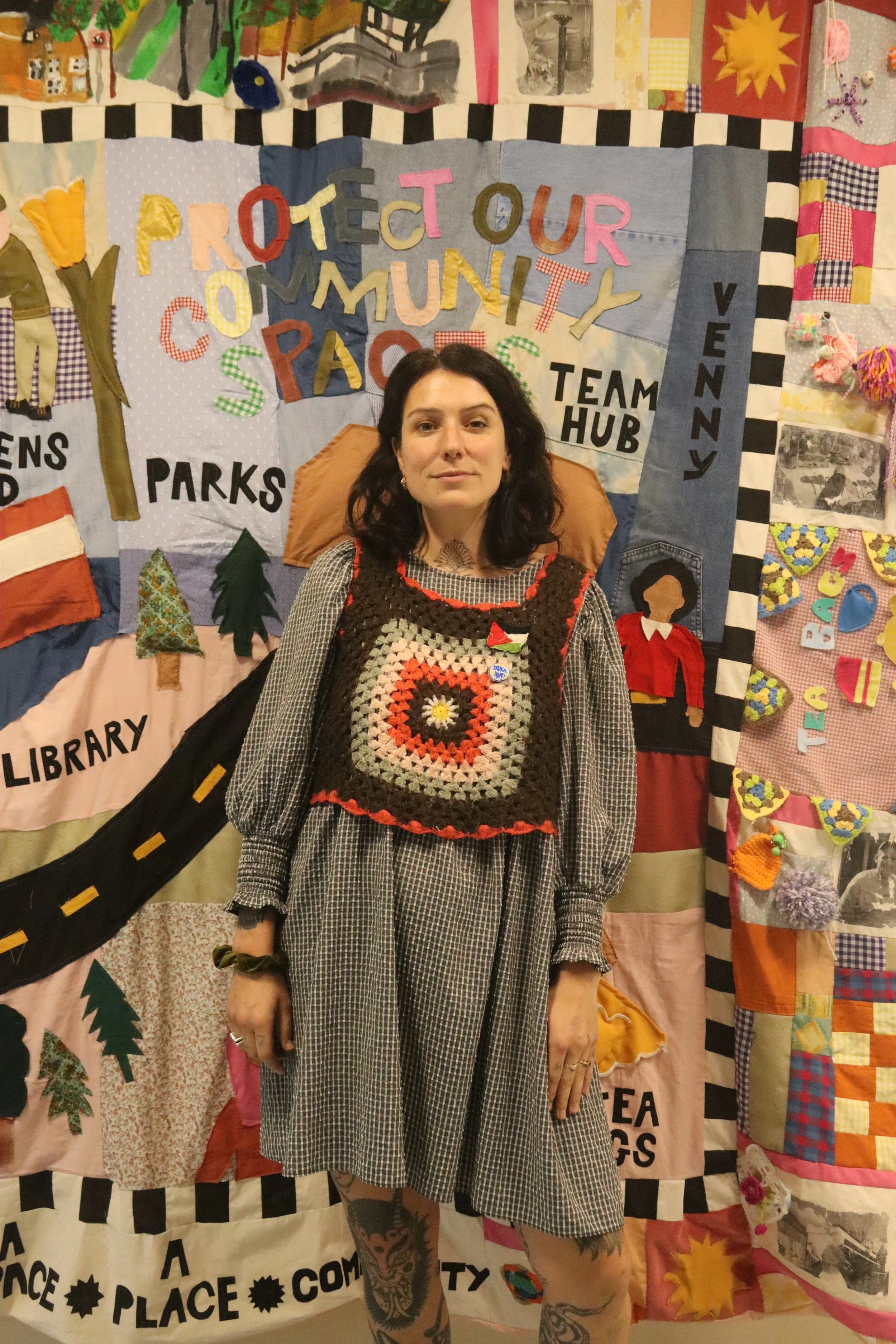 A smiling woman with tattoos and dark hair. Bean Mhi stands infront of a patchwork banner