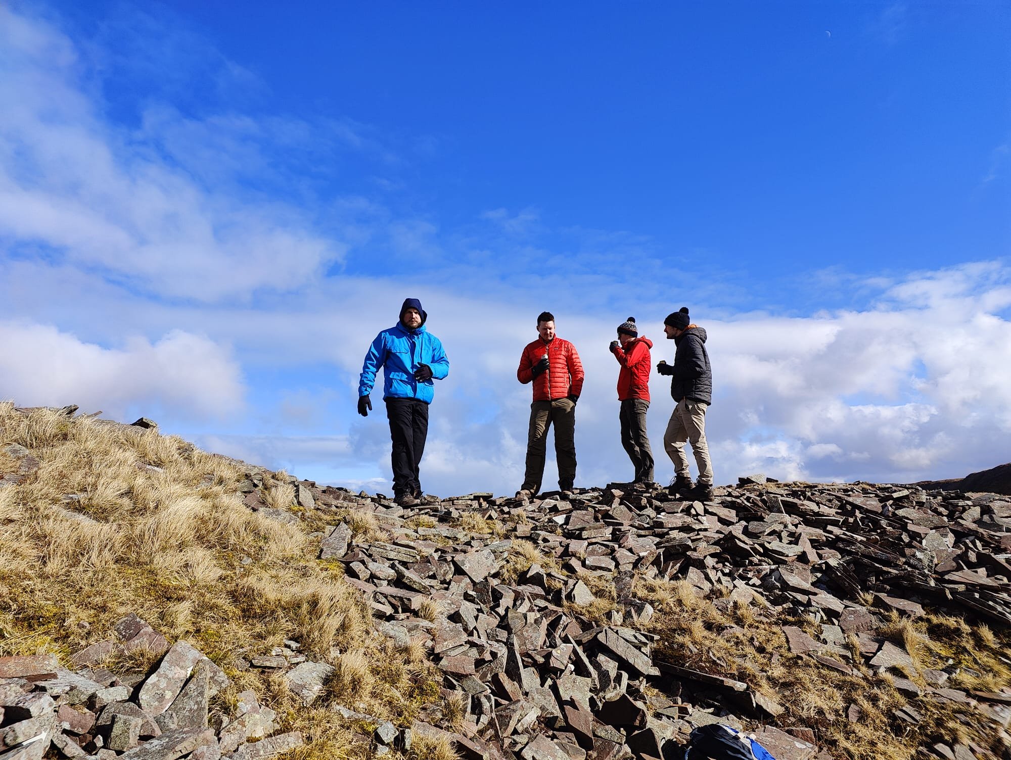   People stood on rocks on top of a hill in warm winter clothes and a drink in hand, blue sky in the background  