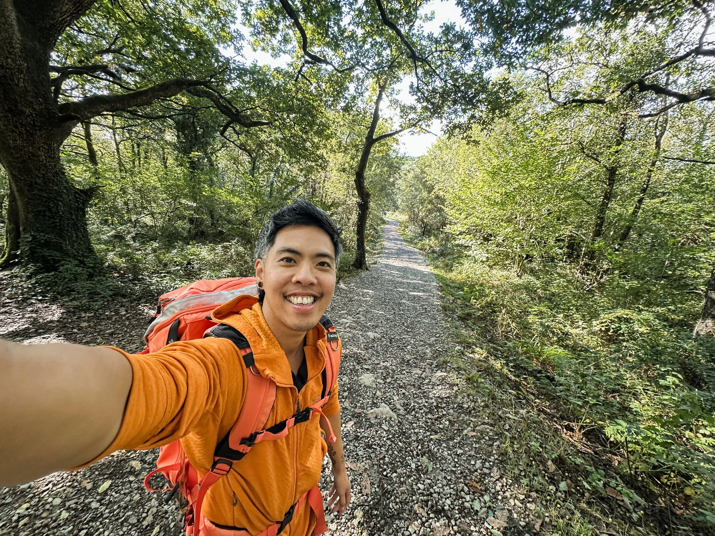 Frit is a Chinese trans man with short black hair, dark brown eyes, sometimes wears glasses. In this photo he is on a woodland path wearing orange and taking a selfie.