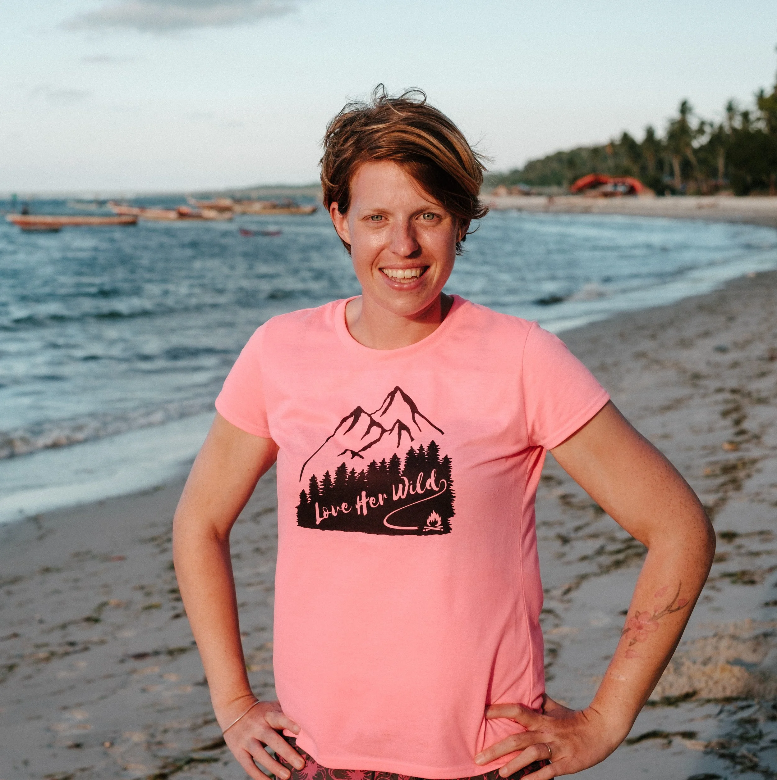 An image of Bex, a white woman with short brown hair wearing a pink t-shirt with love her wild on it. she is stood by the sea