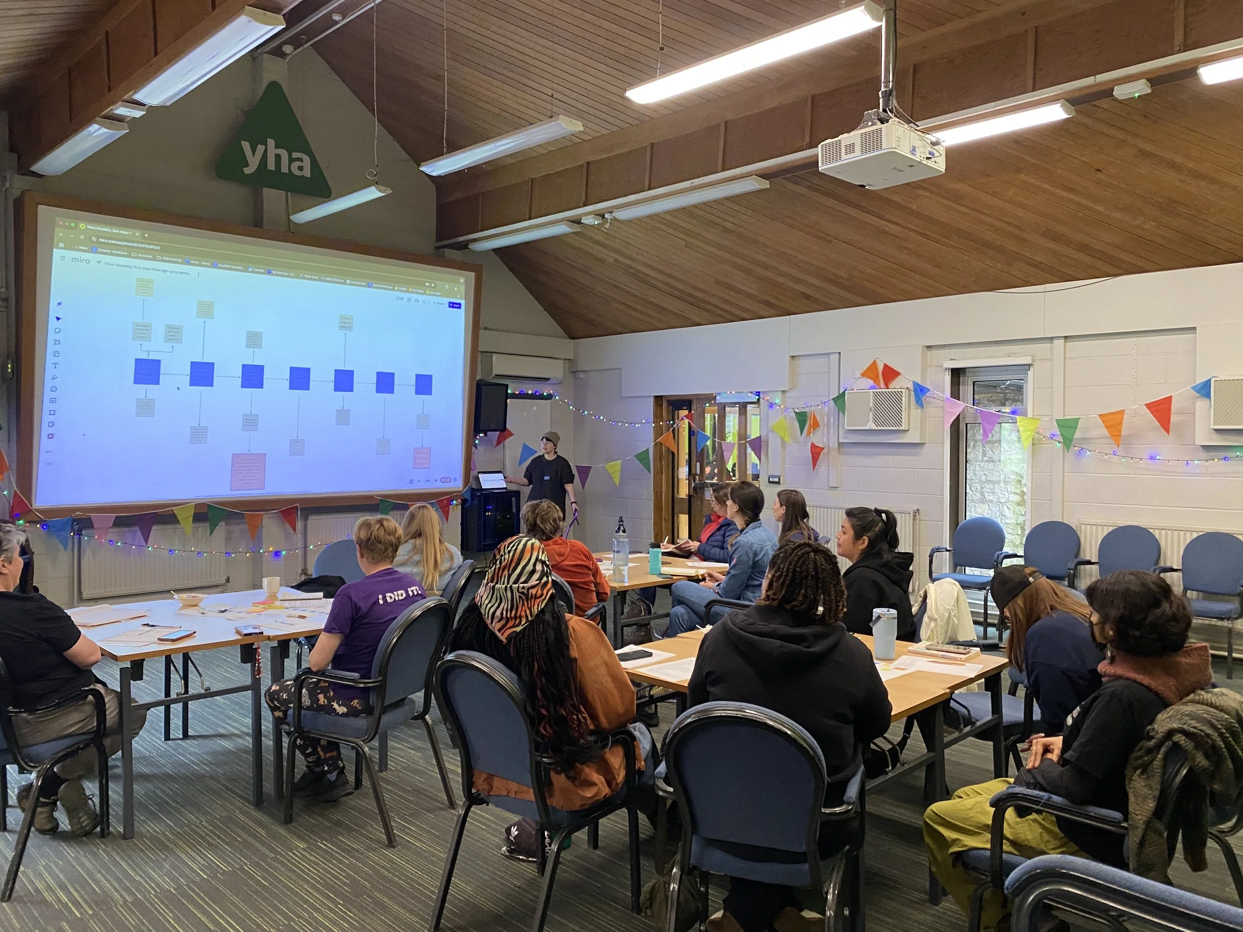 A room full of event attendees, they sit looking at a presentation, Pip and Clare are on the left side of the photo