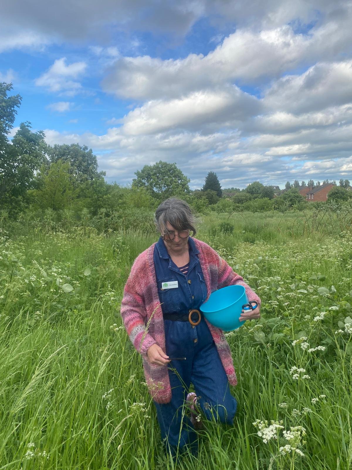 Joss is a white woman in her fifties. She has brown eyes, freckles, and wears glasses. She has shoulder length grey hair. She is standing in a field full of plants and is carrying a blue bowl and a pair of scissors