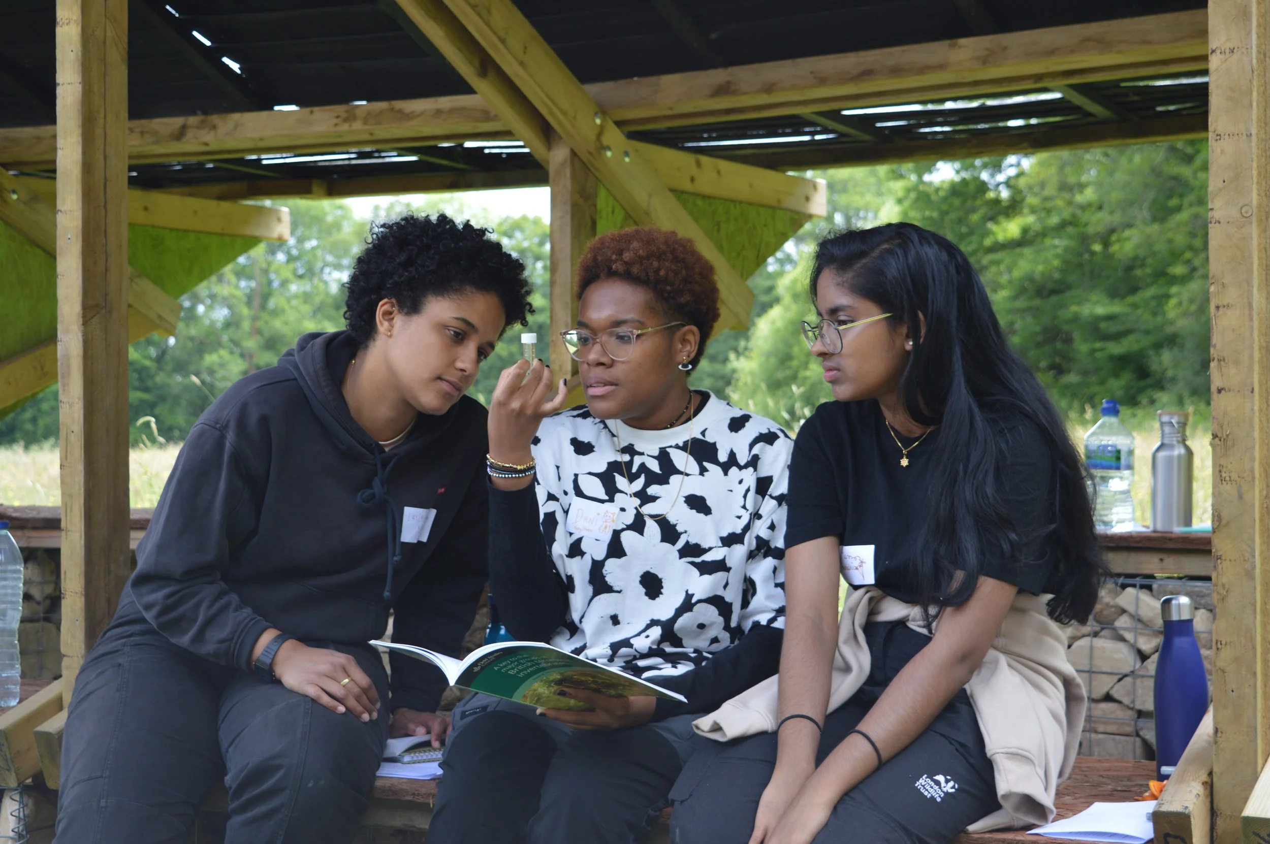 Three people of colour sit with a book open and sample in hand - they are in a shelter but there is a rich green woodland behind them.