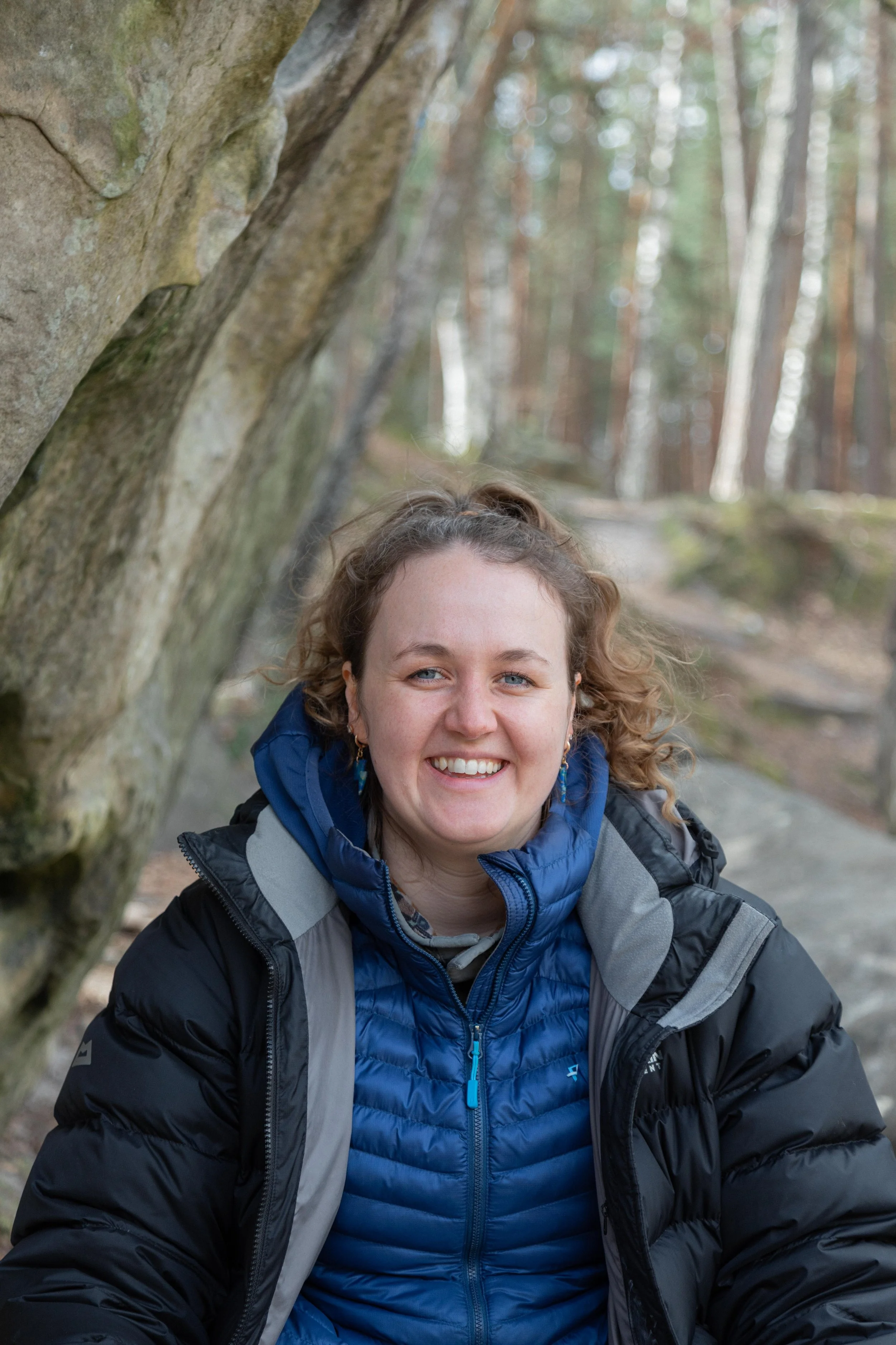 Cisgender, white, woman with blondeish hair, blue eyes, size 12 ish and 170cm tall. Emily sits infront of a boulder in a forest.