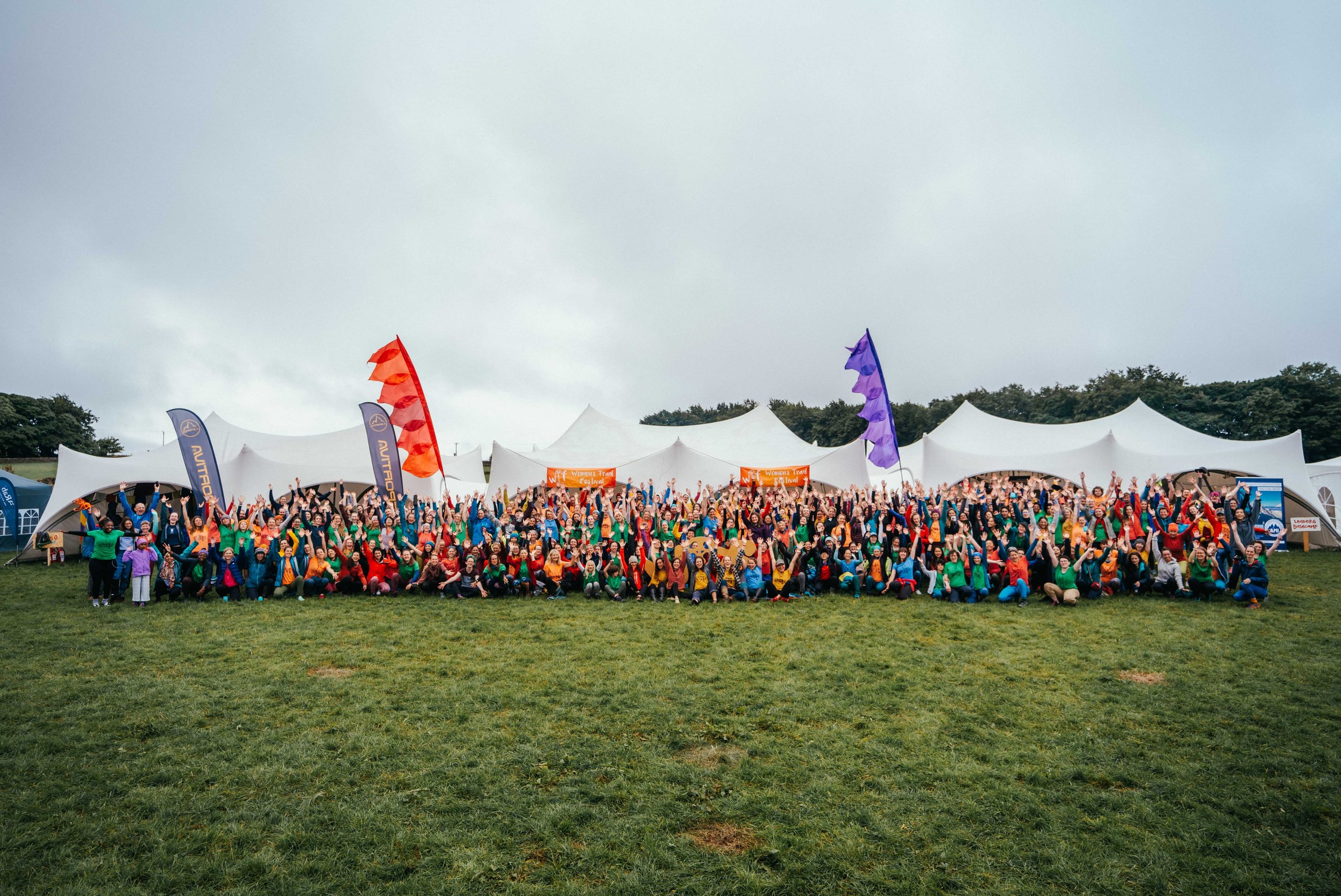 A wide angle group photo of hundreds of women in red, orange, green blue tshirts there are flags and large tents behind them.