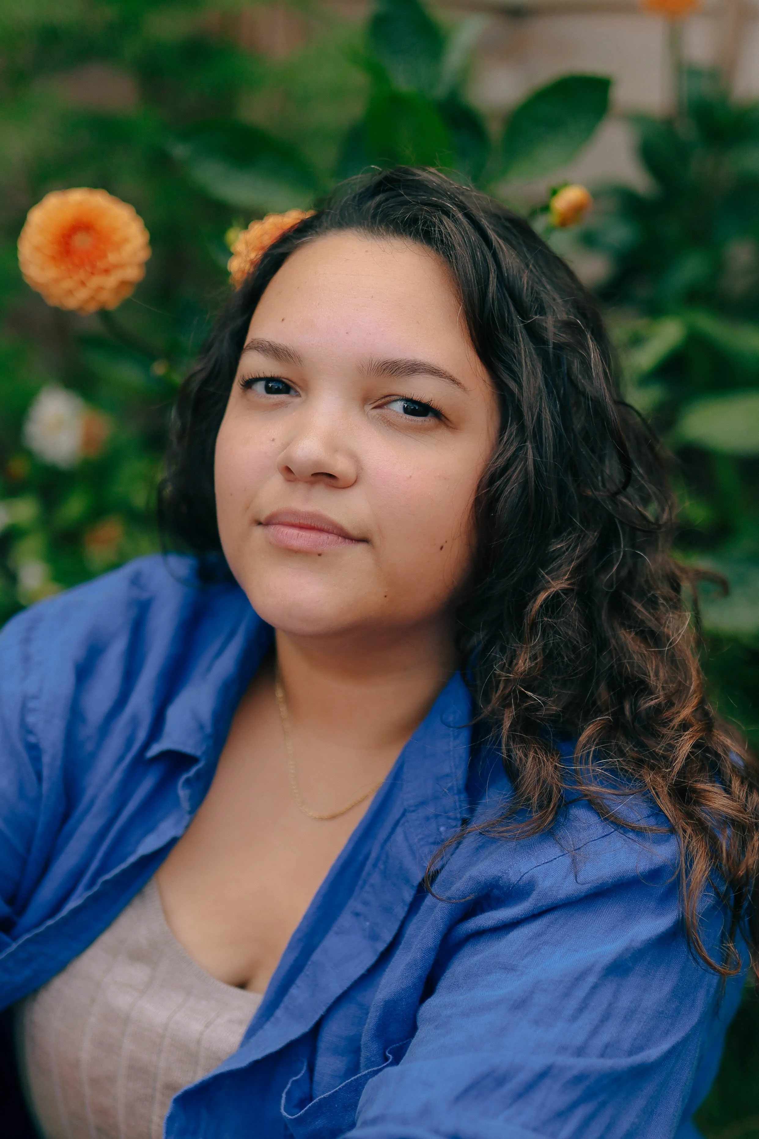 Celie is a mixed heritage woman with long brown, curly hair and dark brown eyes. In this photo she sits in front of flowers looking at the camera.