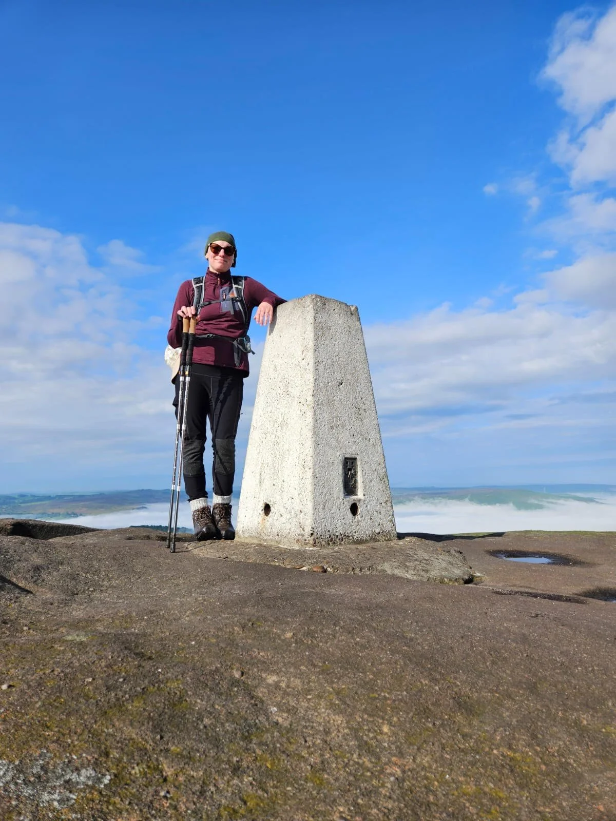 Dawn stood by a trig point in walking gear next to the coast