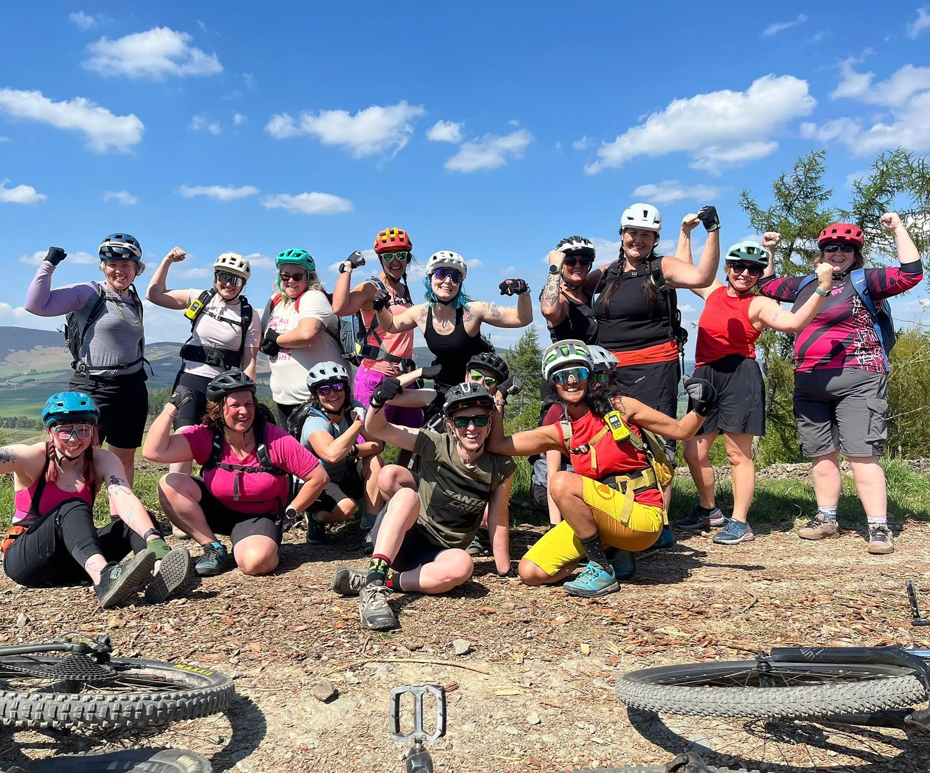A group ow women in MTB gear flex their arms - there are MTB wheels in the front of the photo