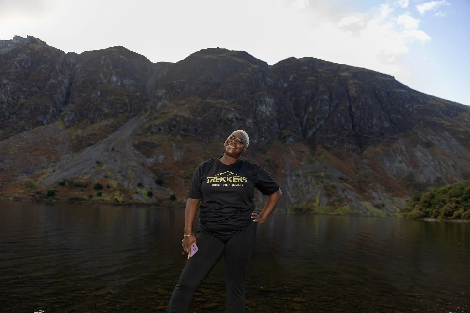 Monique a blck woman with bright white har stands wearing a black Trekkers t-shirt infront of a mountain lake