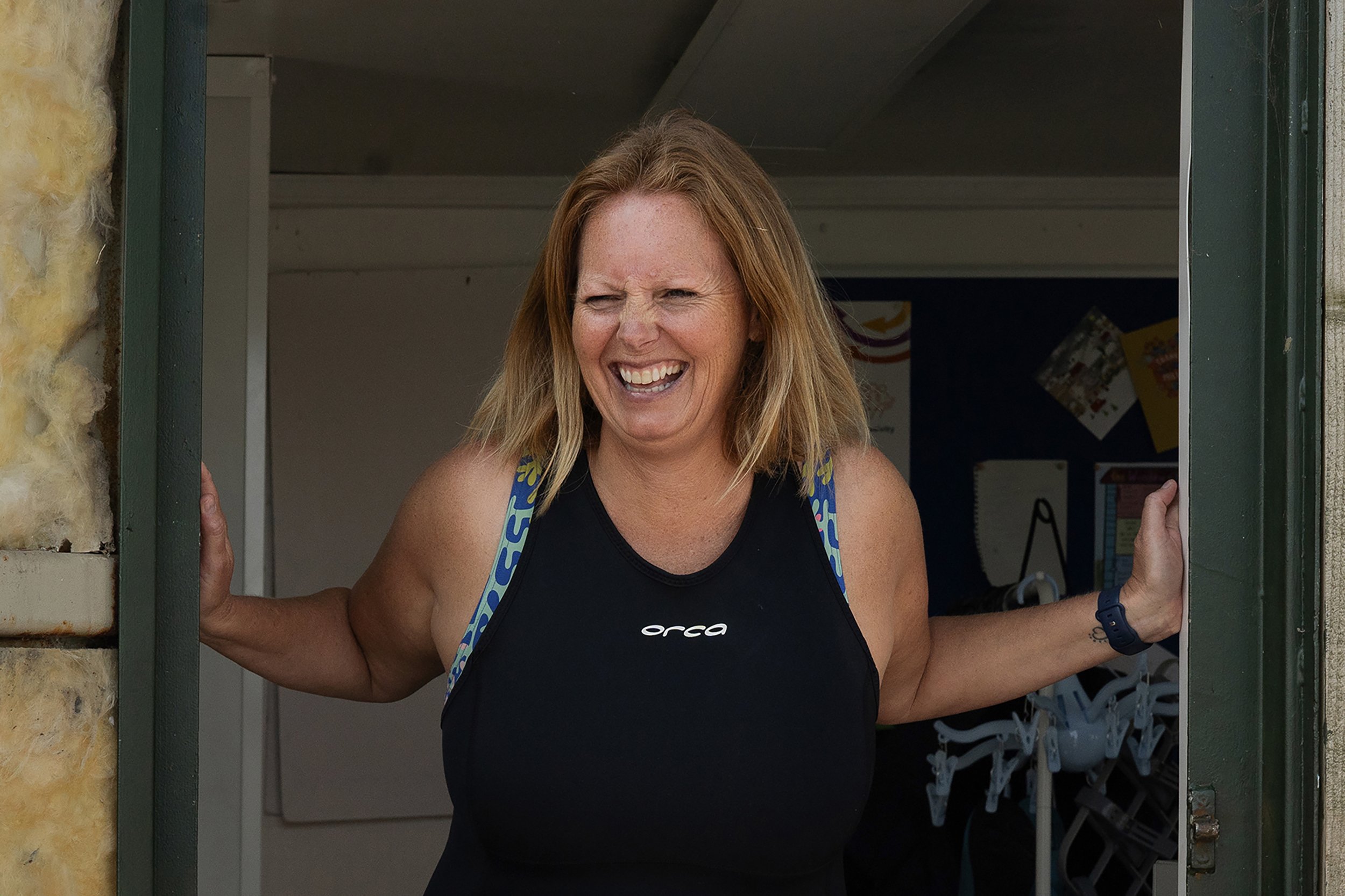 Maggie, a white, tall woman with auburn hair in a chin length bob, blue eyes leans through a doorway smiling.