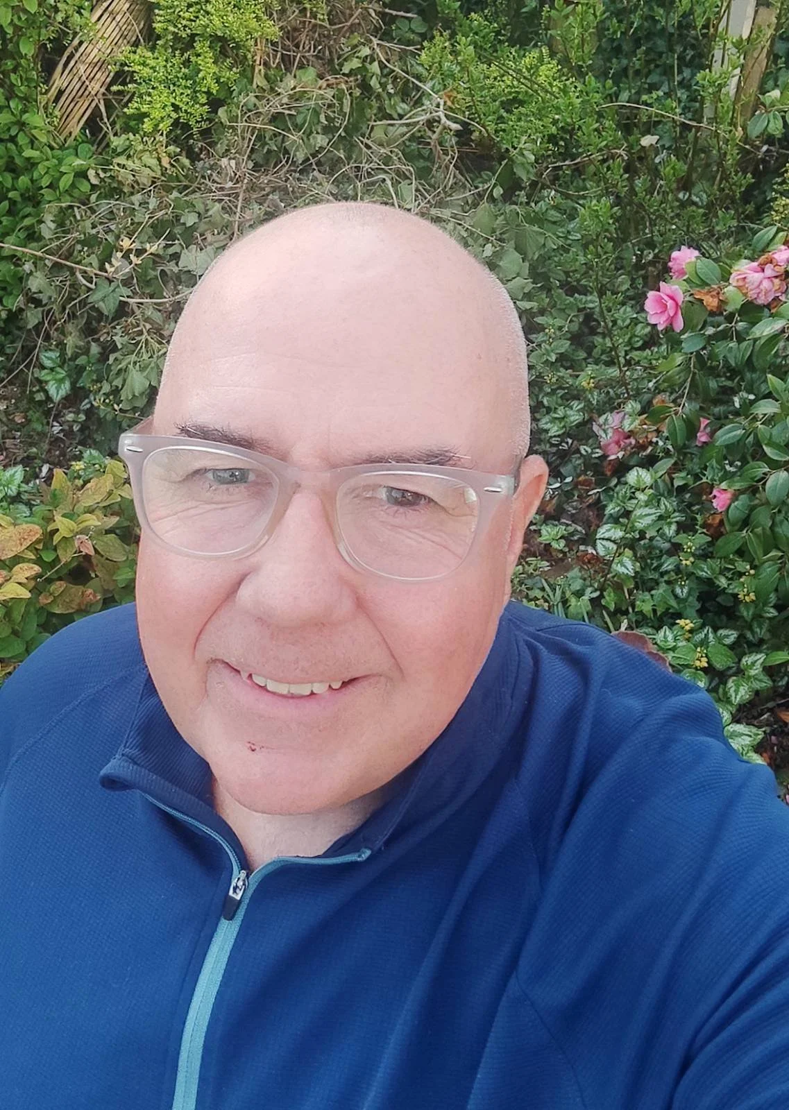 Mike Bird, is male, white British, with short grey hair, brown eyes. In this photo he smiles at the camera with colourful pink flowers behind him.