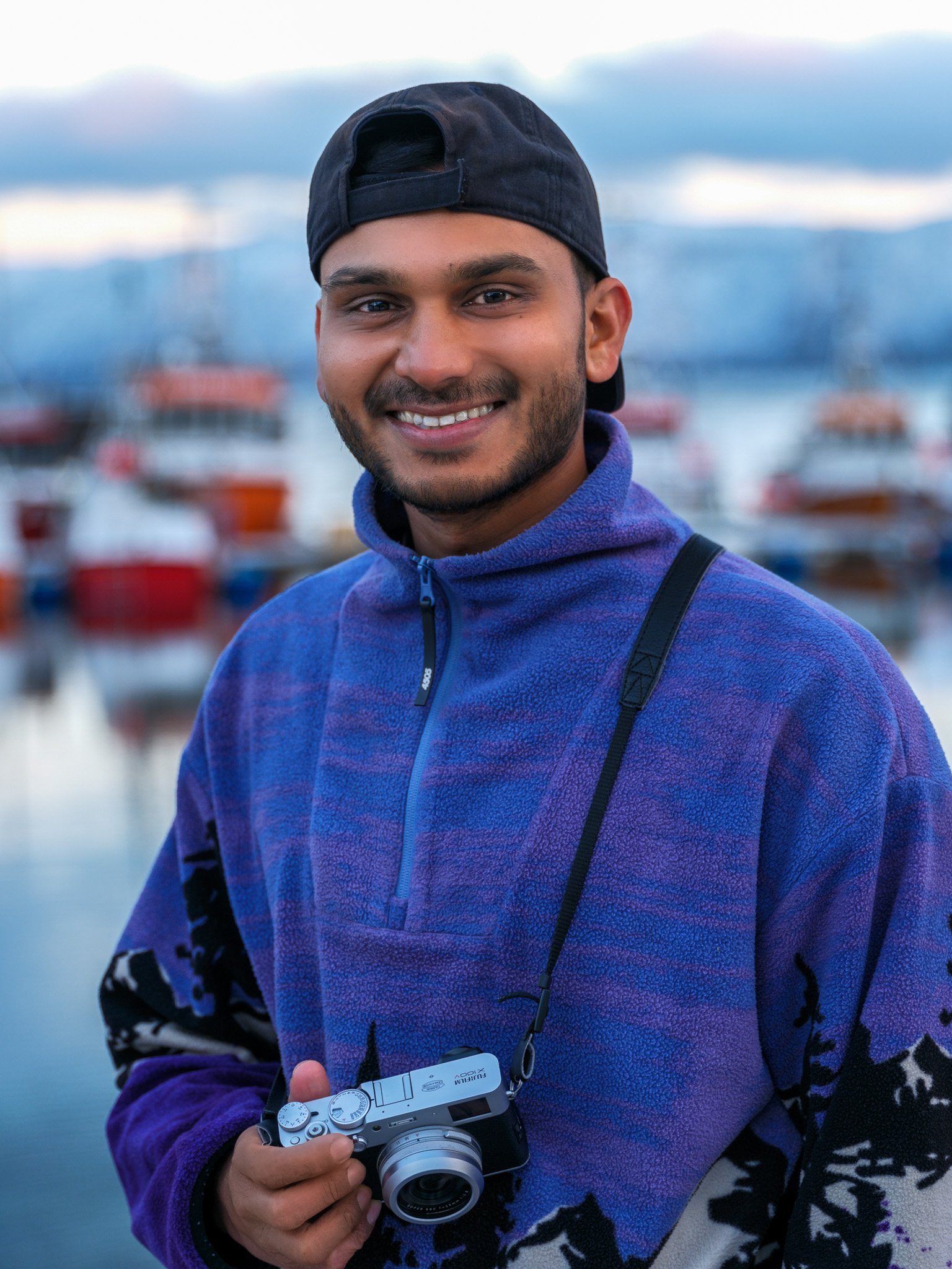 Male, Indian, Black hair, brown eyes, 5 foot 10. Les wears a purple fleece holding a small camera, they are stood in front of a blurry port.
