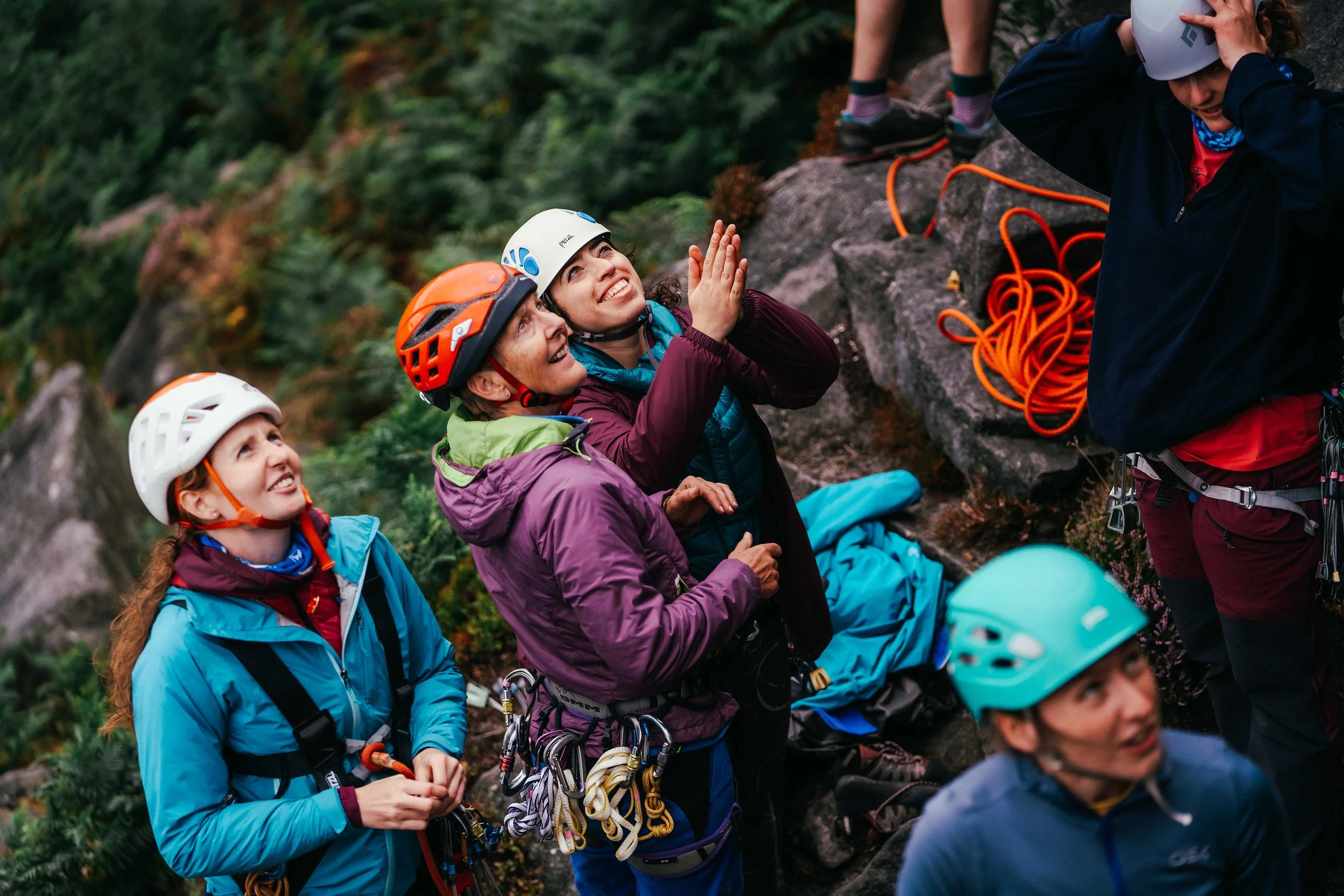 A group of women look up at a climb there is trad gear around them and they are all smiling and clapping