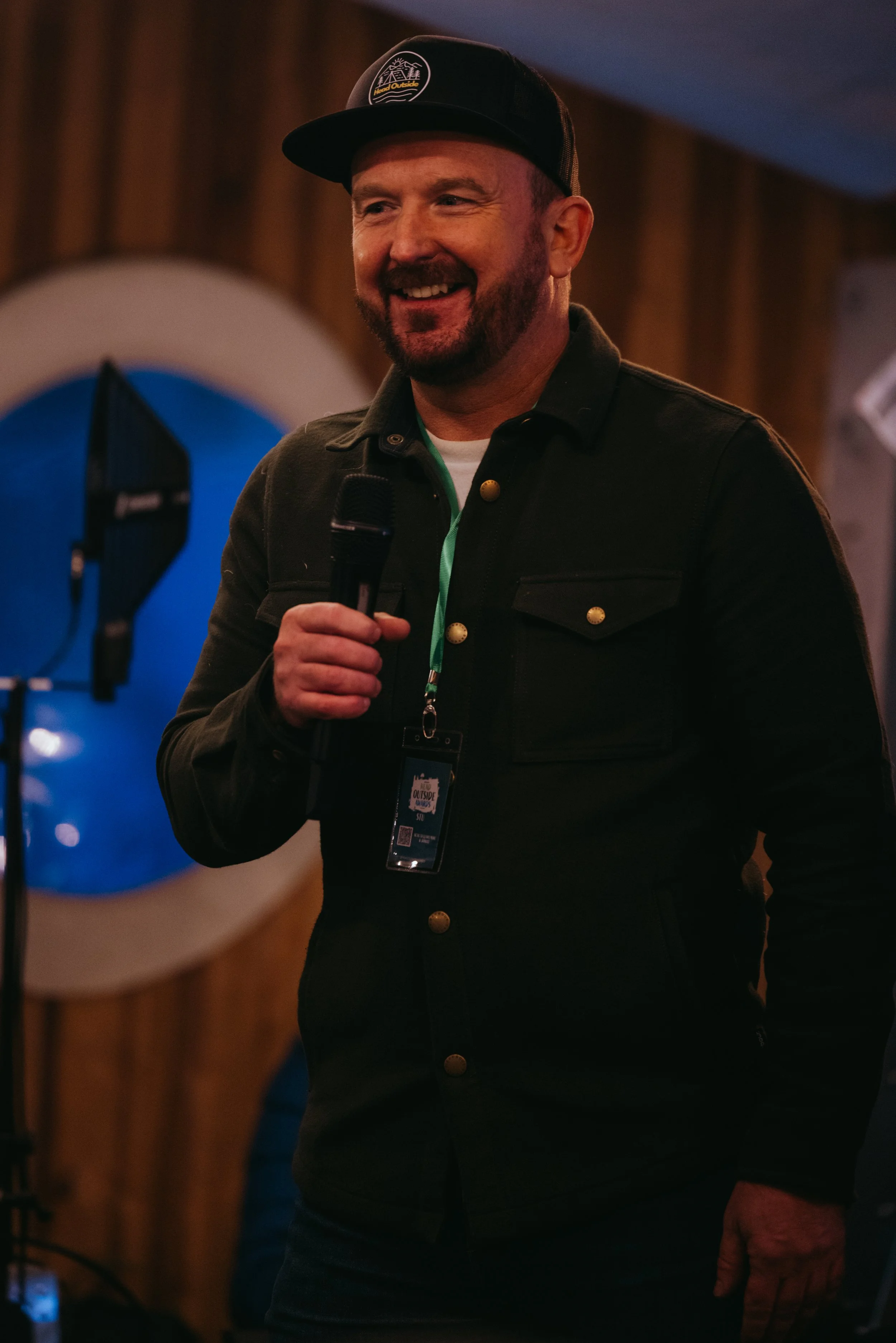 Stu is a white man with a brown beard. In this photo he is wearing a black hat and talking into a microphone.