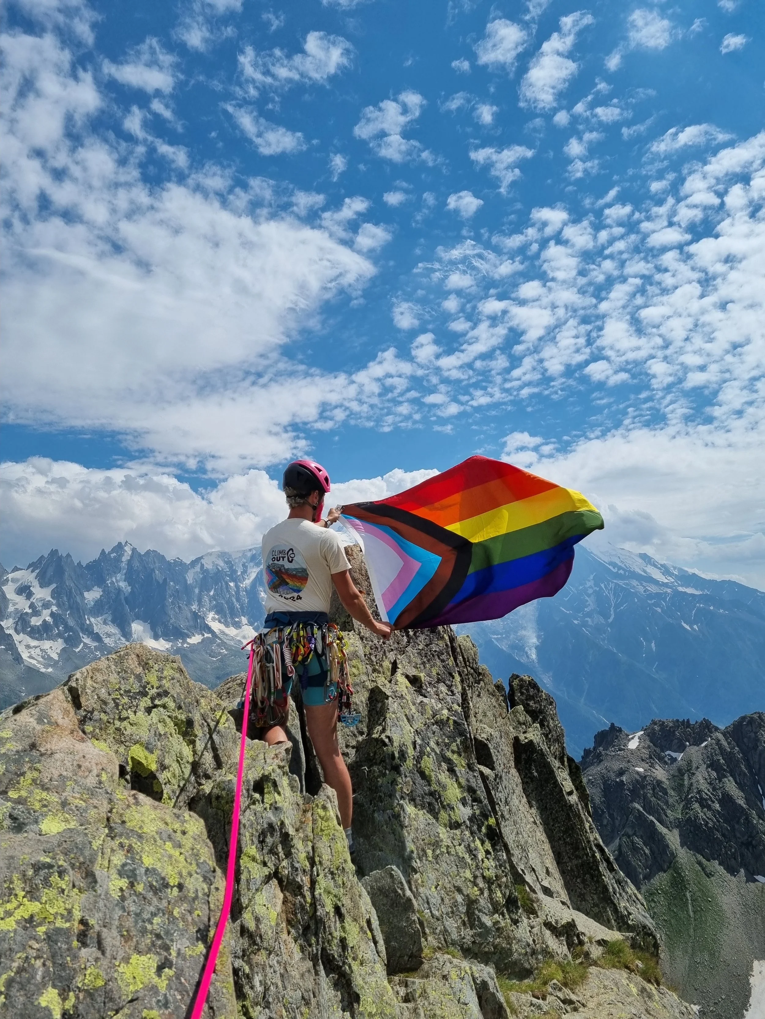 AJ is in the mountains with a full climbing rack, a ClimbOut tshirt and a Pride progress flag.