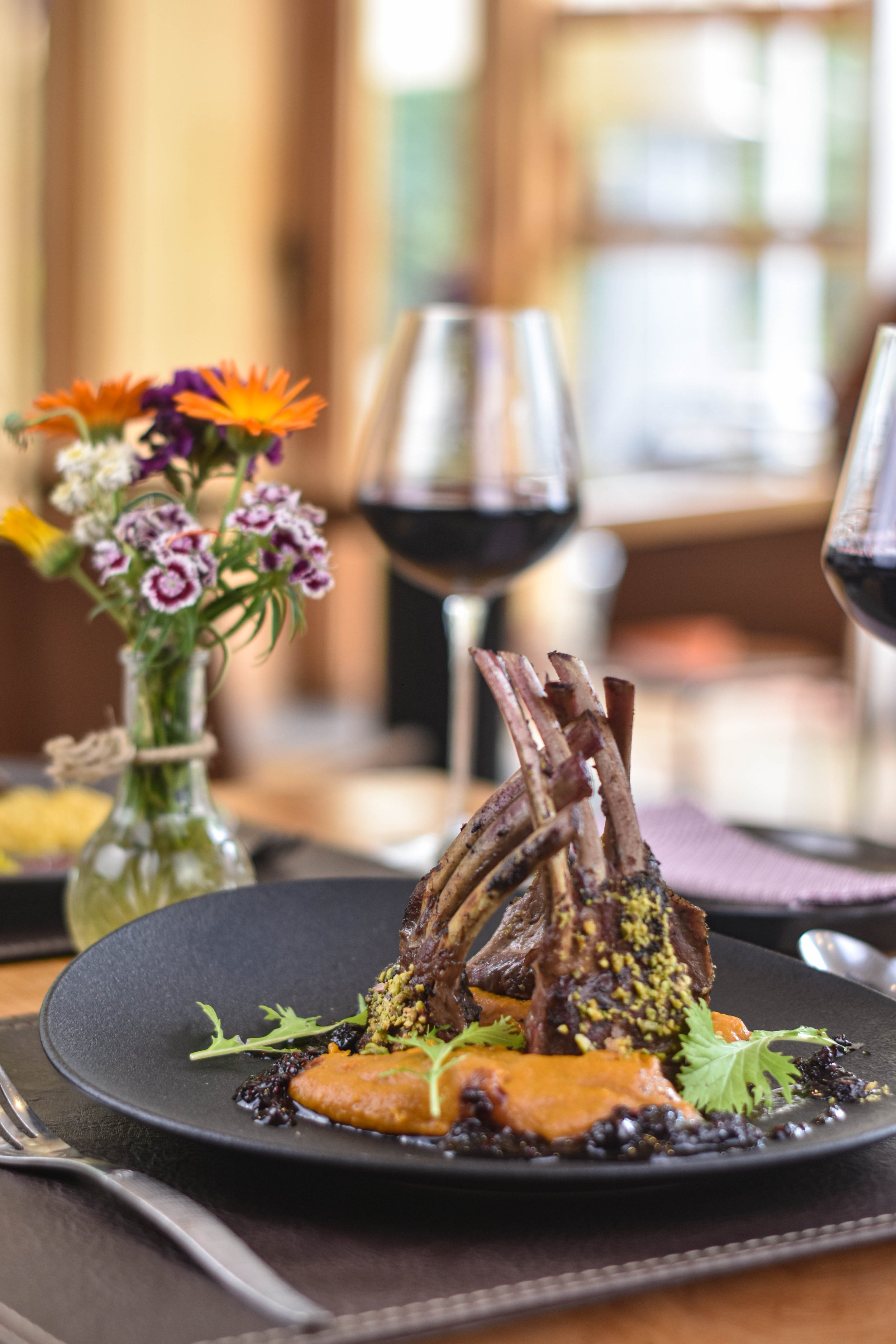 Plato con costillas de carne, puré de un color anaranjado y verduras verdes en un plato negro. En el fondo hay una copa de vino tinto, un jarrón con flores y una ventana que deja entrar luz natural.
