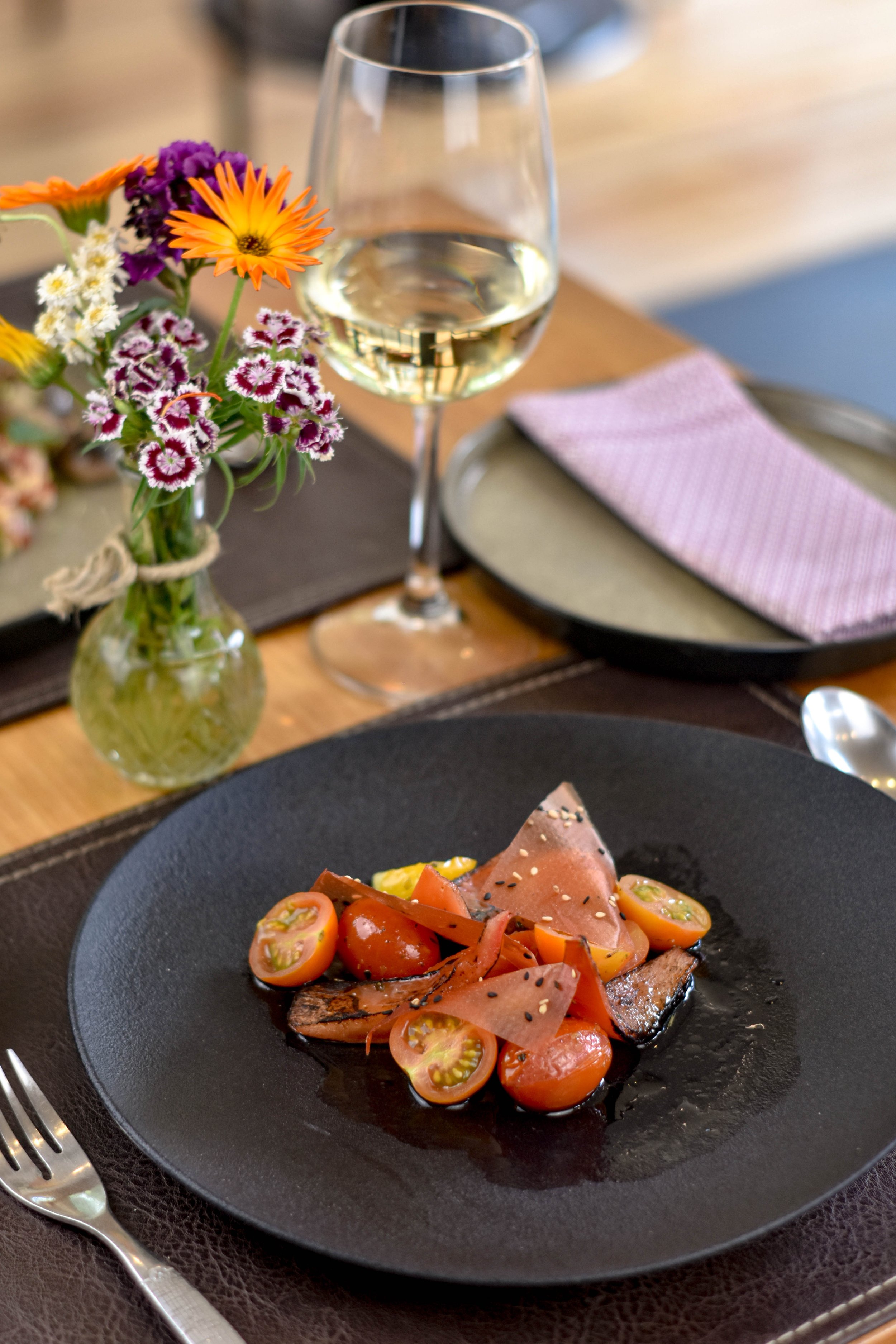 Plato de comida con tomates cherry, rebanadas de carne y salsas, vaso de vino blanco y un florero con flores de colores en la mesa