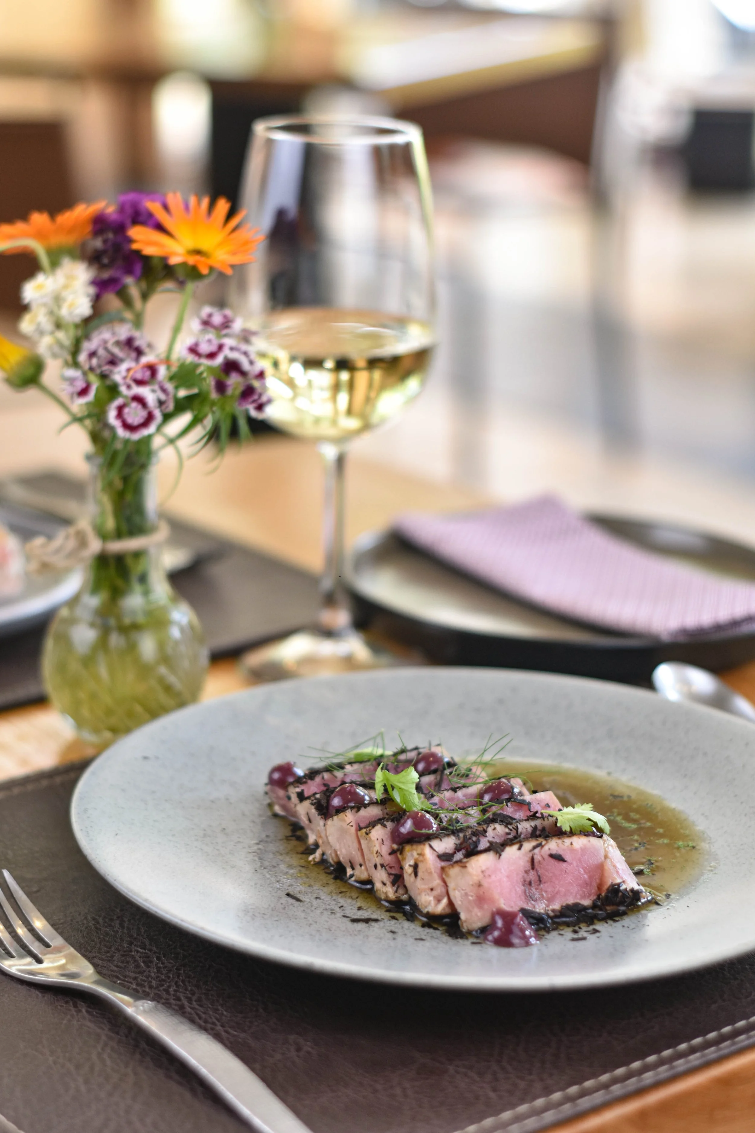 Un plato de atún sellado con salsa y adornado con verduras, acompañado de una copa de vino blanco y un jarrón con flores coloridas en una mesa de comedor.