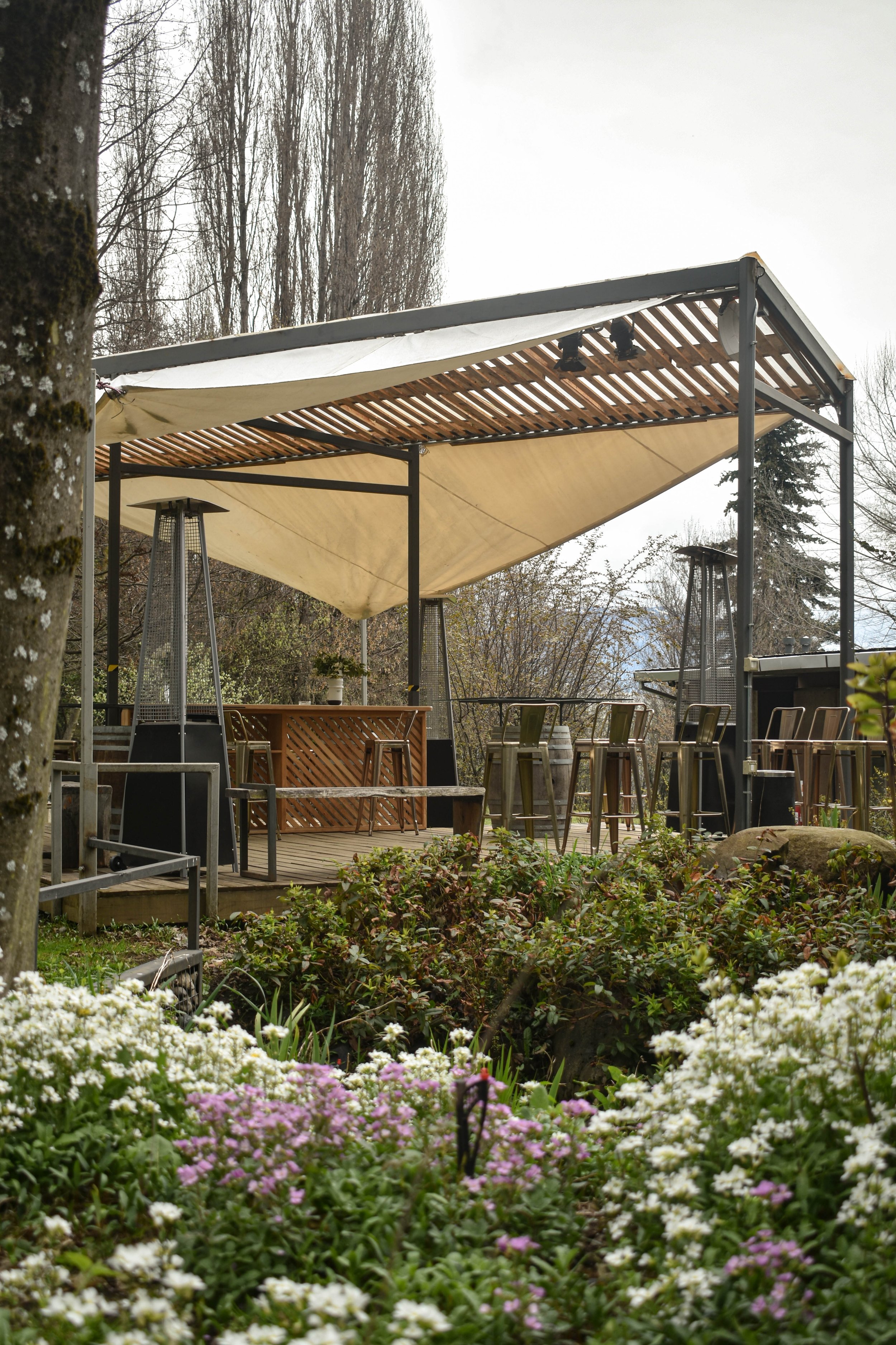 Una terraza al aire libre con un toldo beige, mesas y sillas, rodeada de plantas y flores.