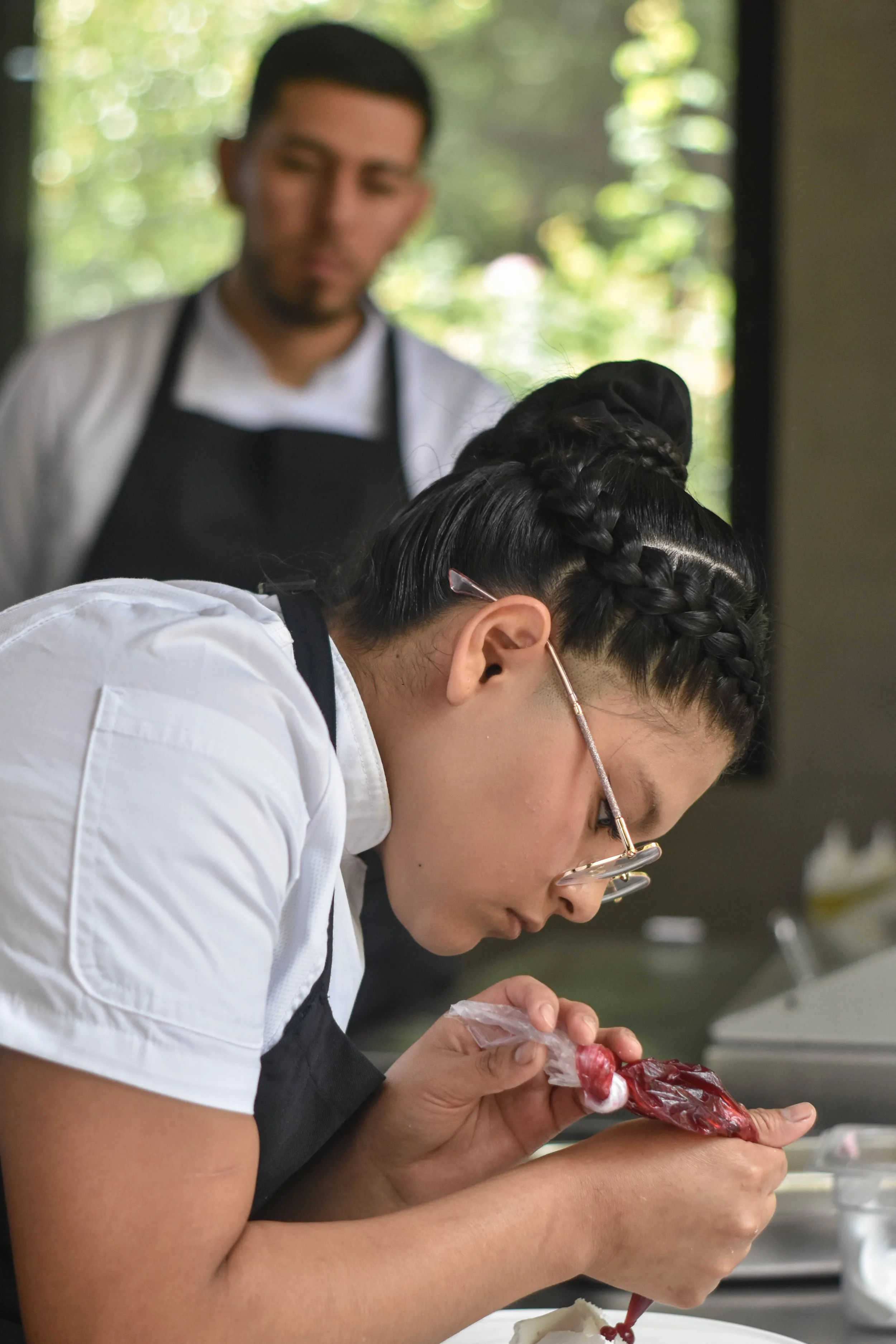Una joven chef decorando un postre con precisión mientras un hombre observa en el fondo, en una cocina moderna con ventanas y vista a naturaleza.