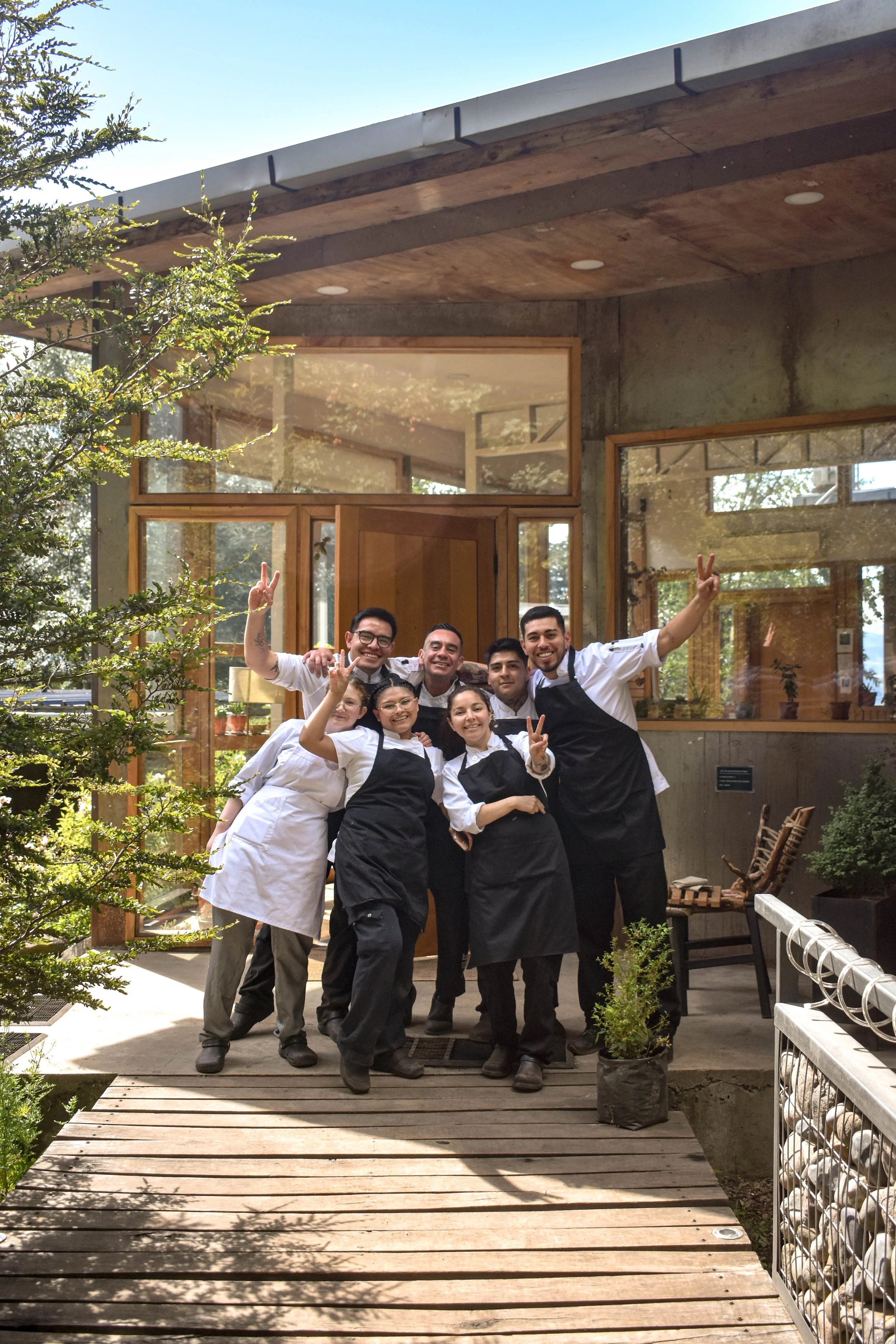 Grupo de siete personas, cocineros y camareros, posando y sonriendo en la entrada de un restaurante con estructura de cemento y madera, rodeados de plantas y árboles.