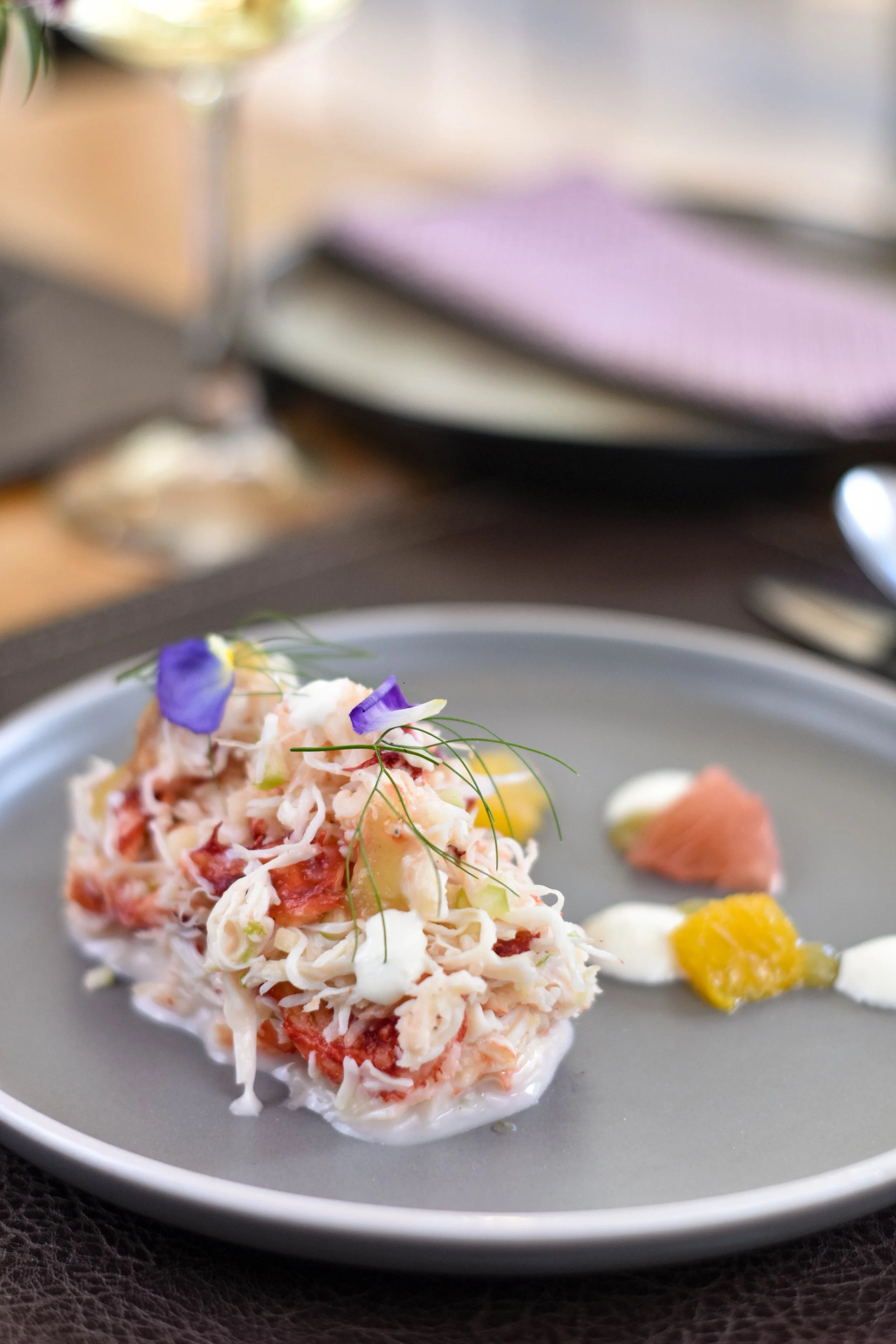En un plato gris, una ensalada de cangrejo decorada con flores comestibles y hierbas, acompañada de pequeñas porciones de salsas con diferentes colores.