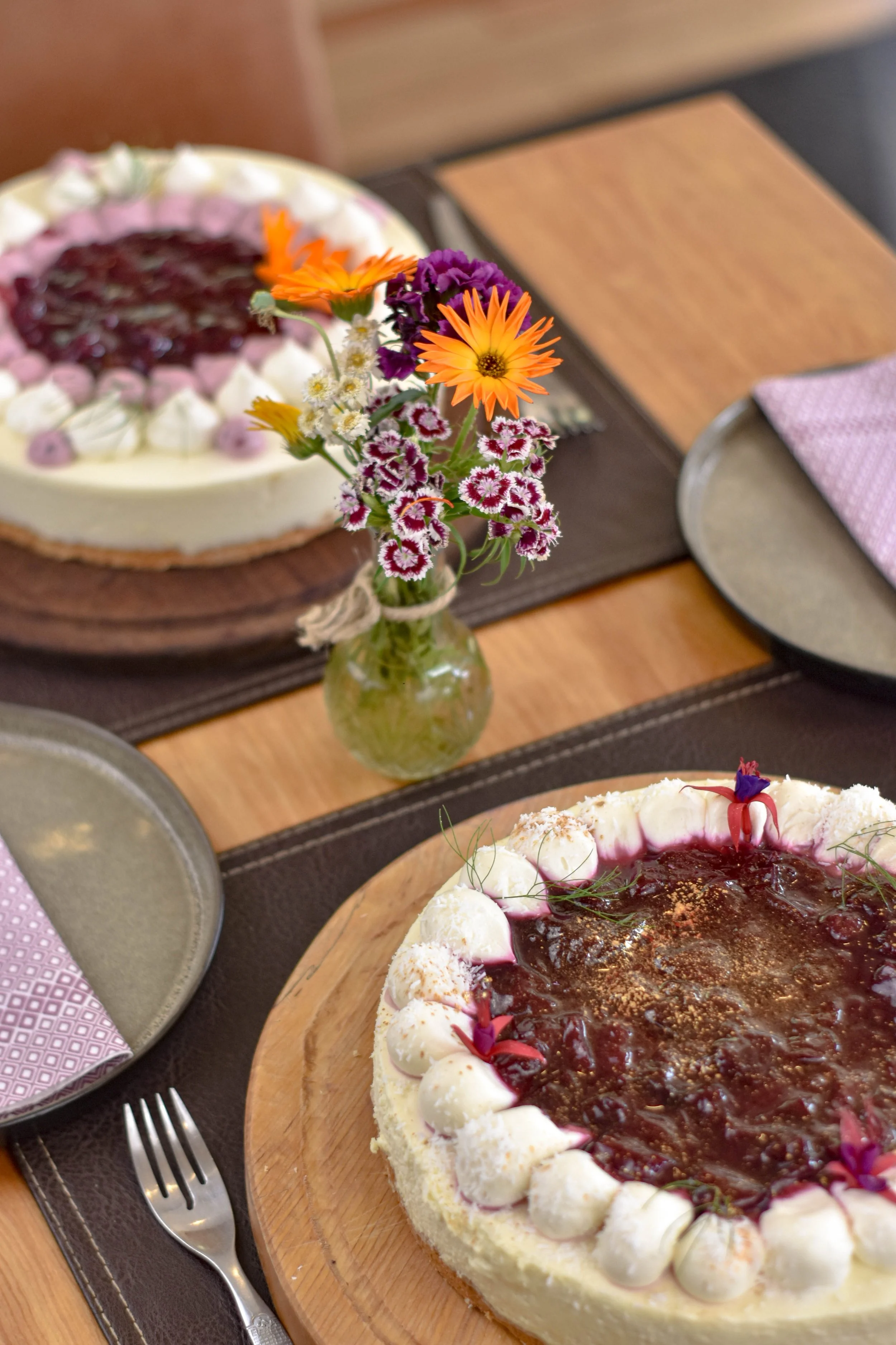 Dos pasteles en una mesa de madera, con un florero con flores coloridas entre ellos.