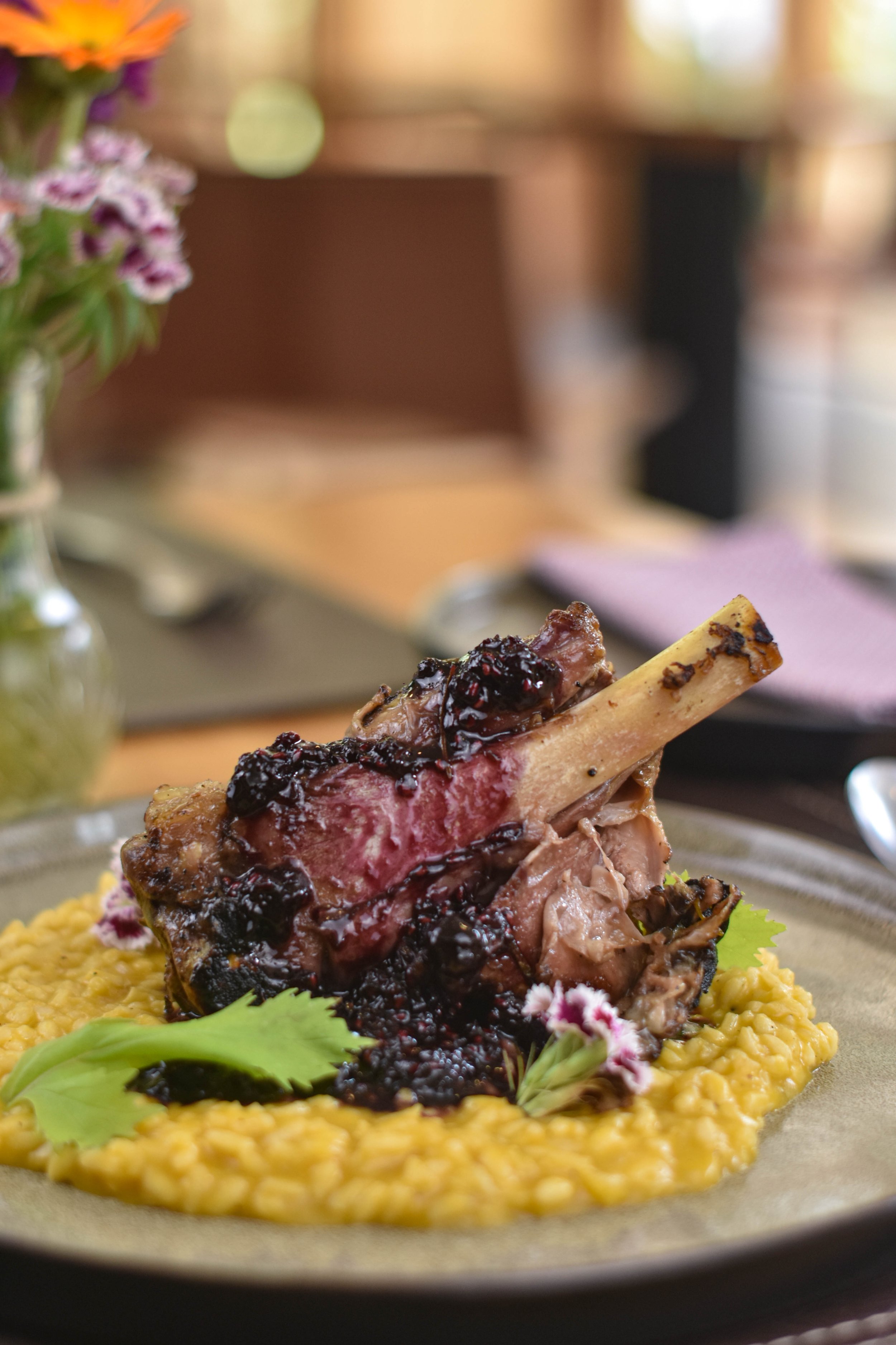 Plato de carne de cordero con salsa de arándanos sobre un puré amarillo, decorado con hojas verdes y flores pequeñas.