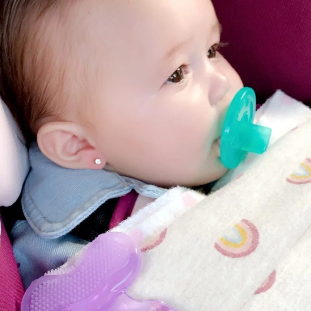 A young child with a pacifier in their mouth, lying in bed, with a rainbow-themed blanket and a purple teething toy beside them.