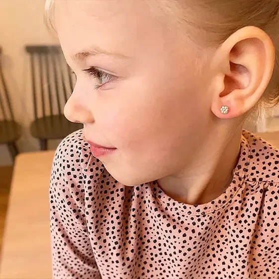 Close-up profile of a young girl with blonde hair wearing flower-shaped earrings and a pink polka-dot shirt, sitting at a wooden table.