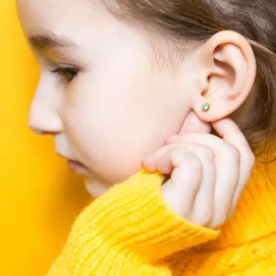 Close-up profile of a young girl wearing a yellow sweater and floral stud earrings, against a yellow background.