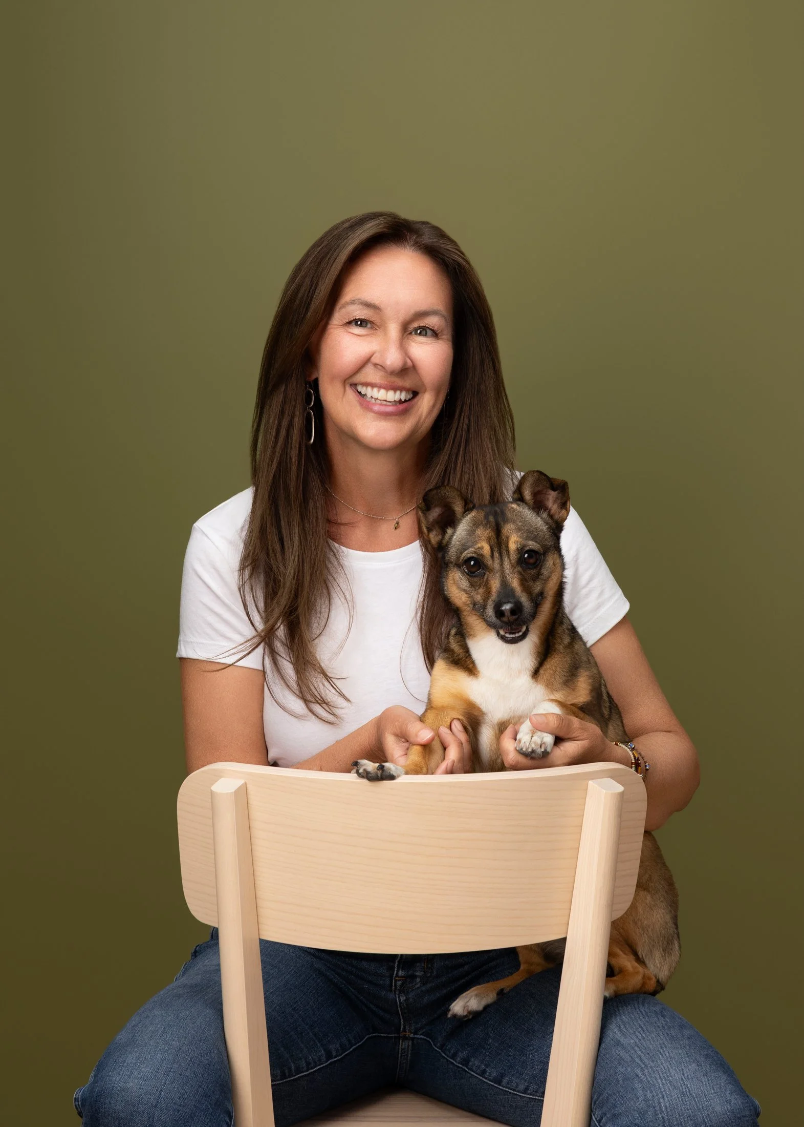 A smiling woman with long brown hair wearing a white t-shirt and blue jeans, sitting on a wooden chair, holding a small brown and black dog with pointed ears and a white chest against a green background.