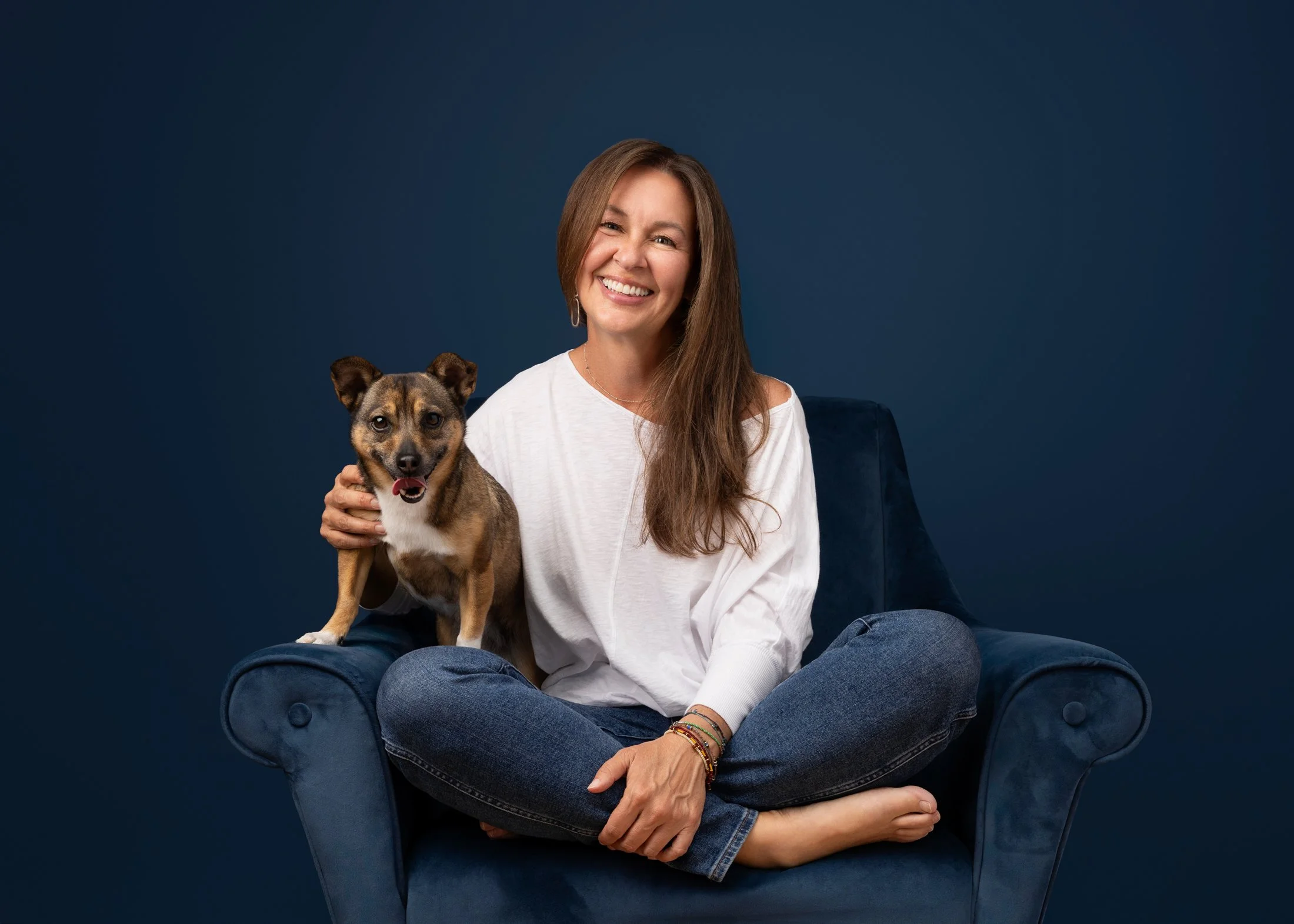 A woman sitting cross-legged on a dark blue armchair, smiling and holding a small dog on her shoulder, against a dark blue background.