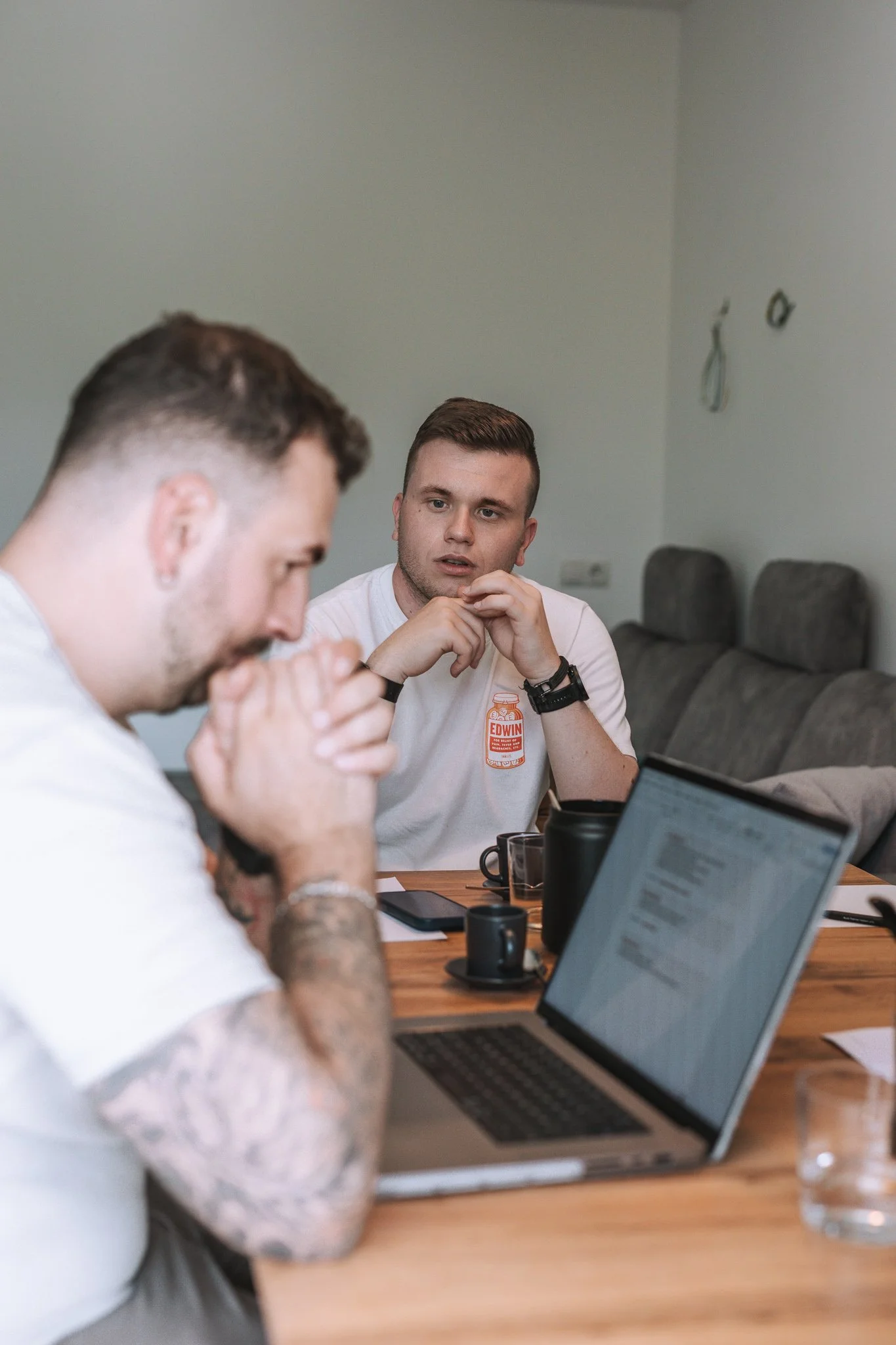 Zwei junge Männer sitzen an einem Tisch in einem Büro und führen eine Diskussion. Der Mann auf der rechten Seite trägt ein weißes T-Shirt mit einem Aufdruck und schaut aufmerksam. Auf dem Tisch befinden sich Laptops, Kaffeetassen und ein Smartphone.