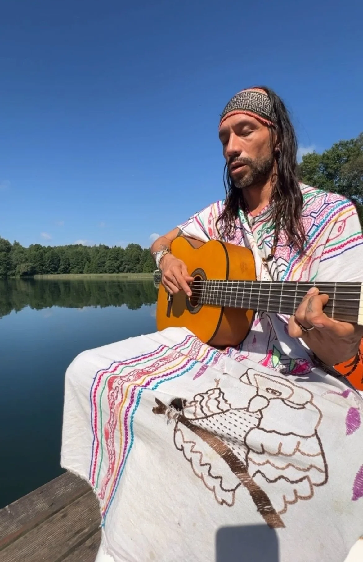 Healer plays a guitar outdoors and heals Icaros near a lake under a clear blue sky. He is seated on a wooden dock, with a colorful embroidered shipibo traditional wear