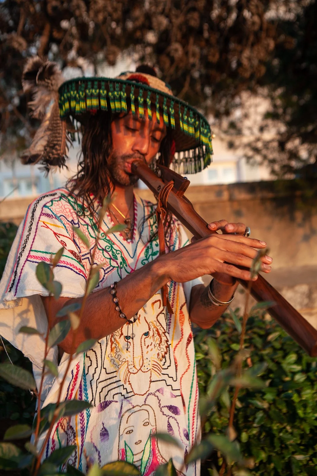 A man wearing a colorful embroidered shirt and a traditional sombrero with feathers, playing a wooden flute outdoors during sunset.