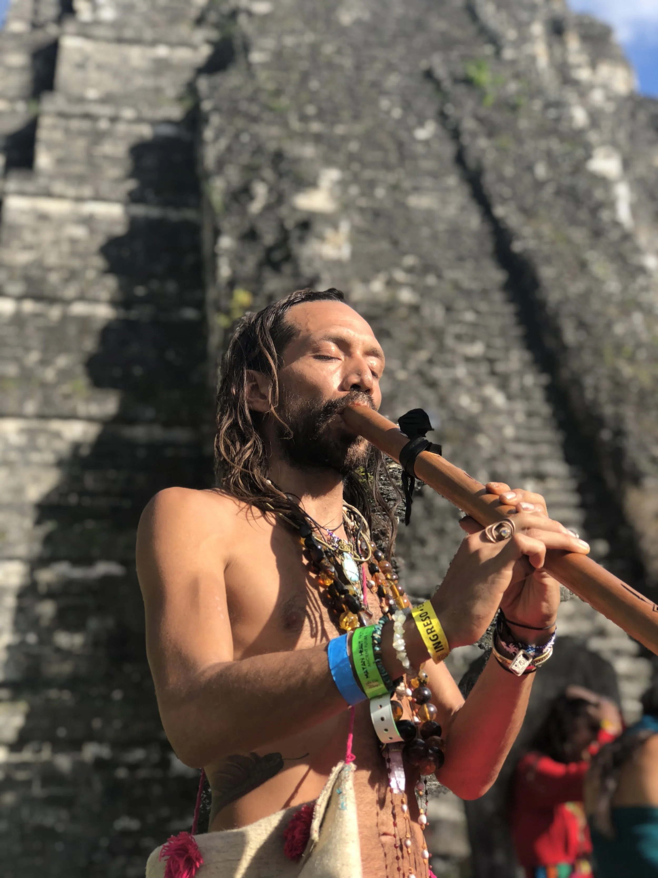A topless man with long hair and jewelry playing a flute outdoors, with a rocky stone structure in the background.