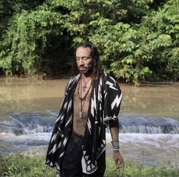 Man with long hair and tattoos, wearing a black and white patterned robe, standing by a river with green trees in the background.