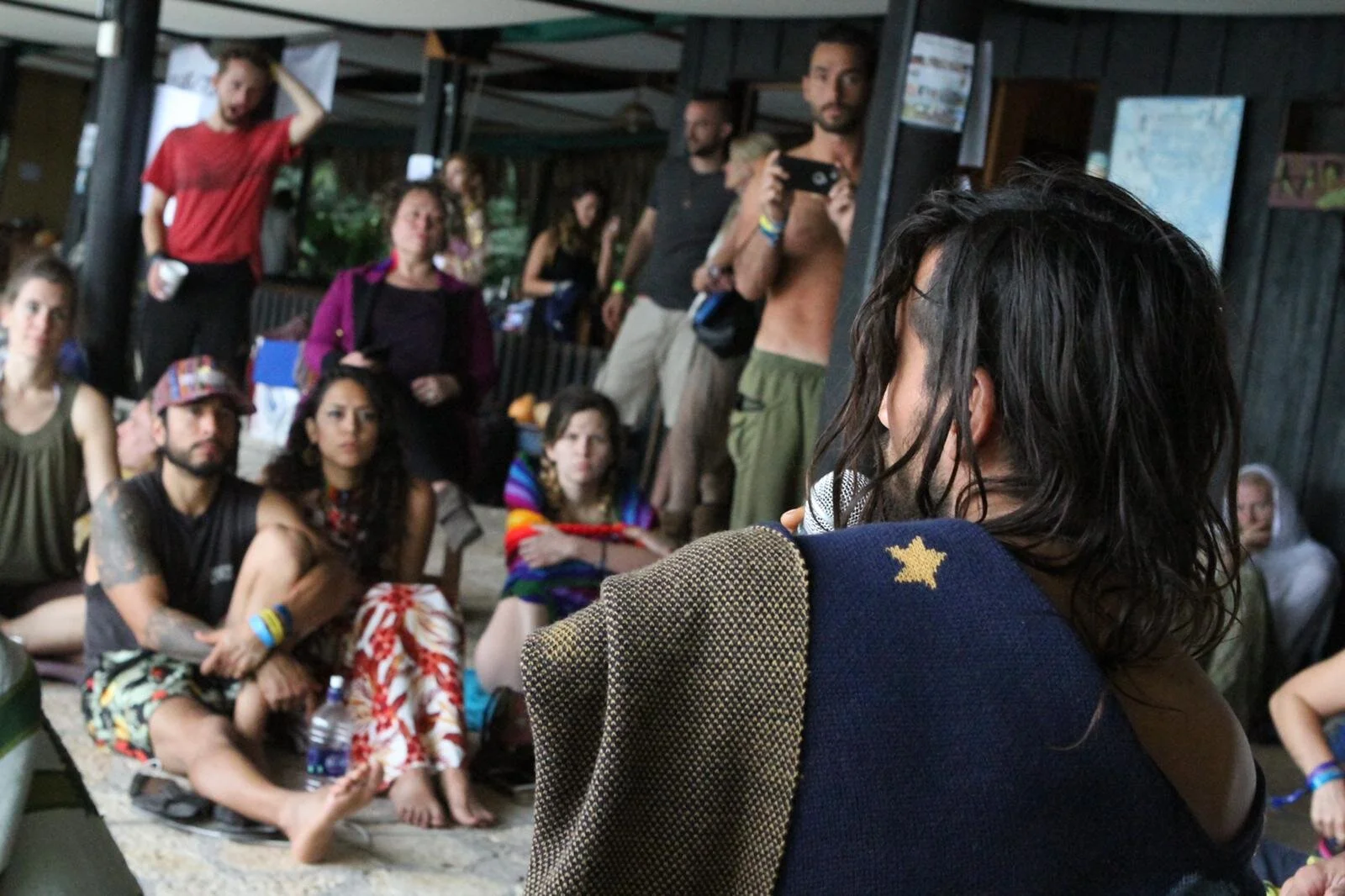 A person with long dark hair, wearing a shirt with a star patch on the shoulder, speaks to a group of diverse people seated and standing, in an indoor setting with dark walls and artwork.