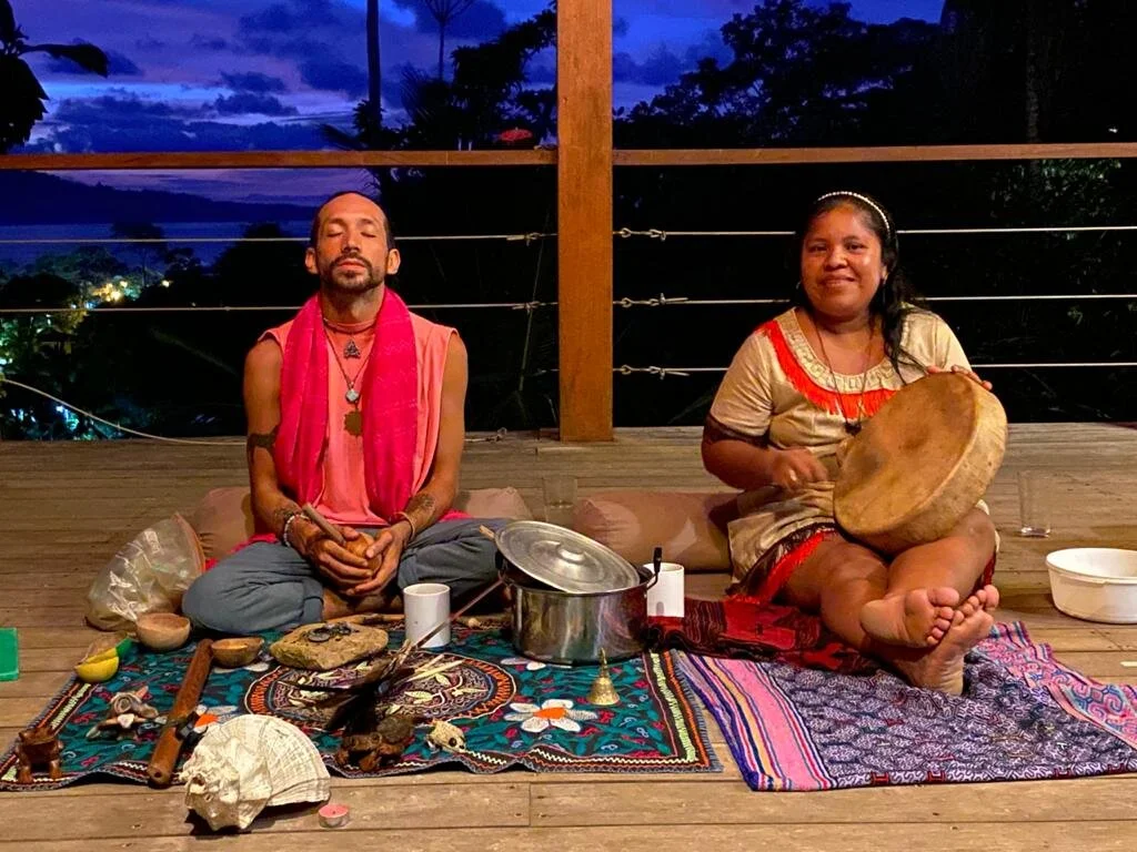 Two people sitting cross-legged on a wooden deck during sunset, surrounded by spiritual objects, with a view of the sky and trees in the background.